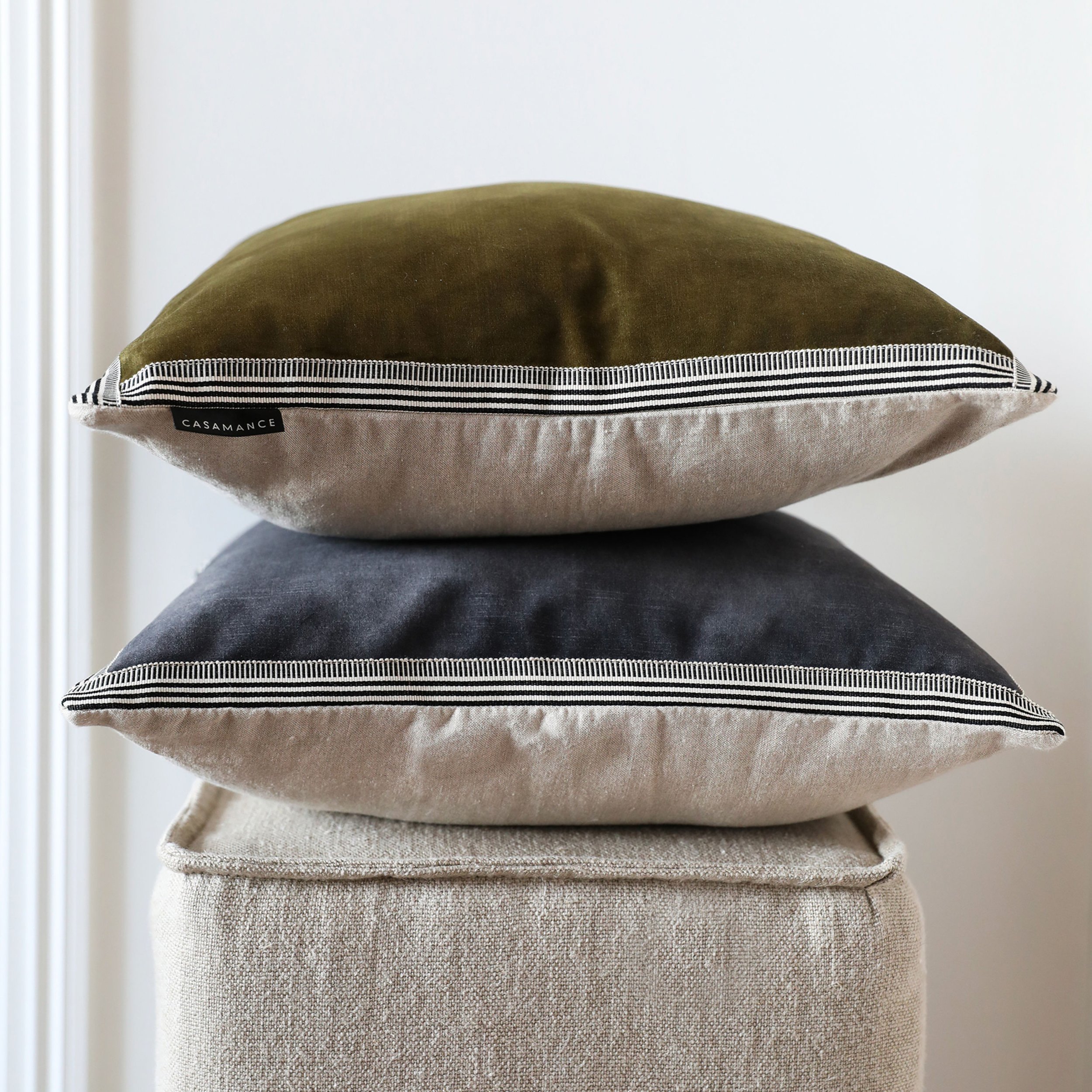 Two Maison Casamance Manade cushions in olive and charcoal stacked on a linen stool, showing the braid edge and linen back