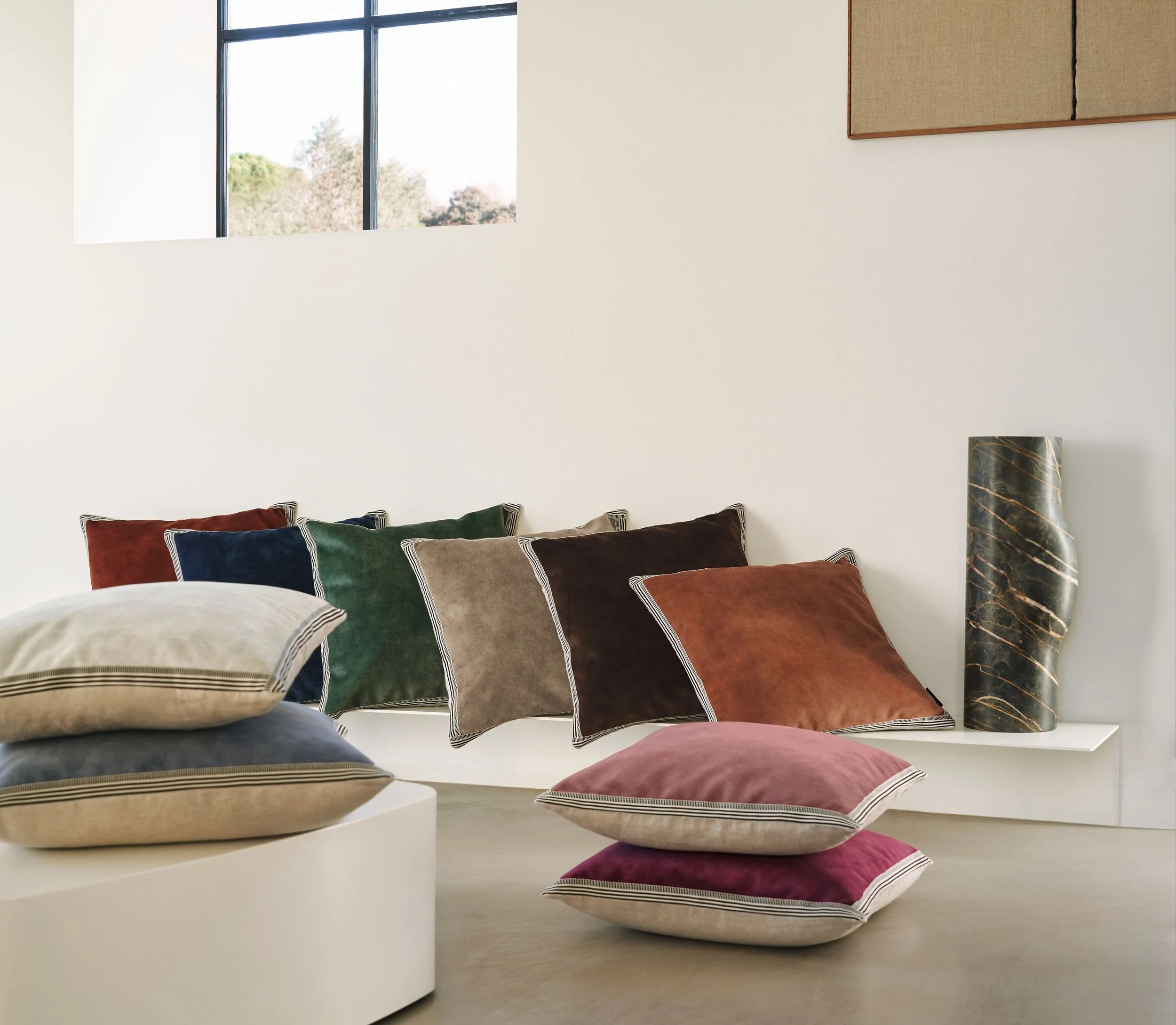 Maison Casamance Manade velvet cushions in rust, navy, green, taupe and pink arranged on a white bench and floor