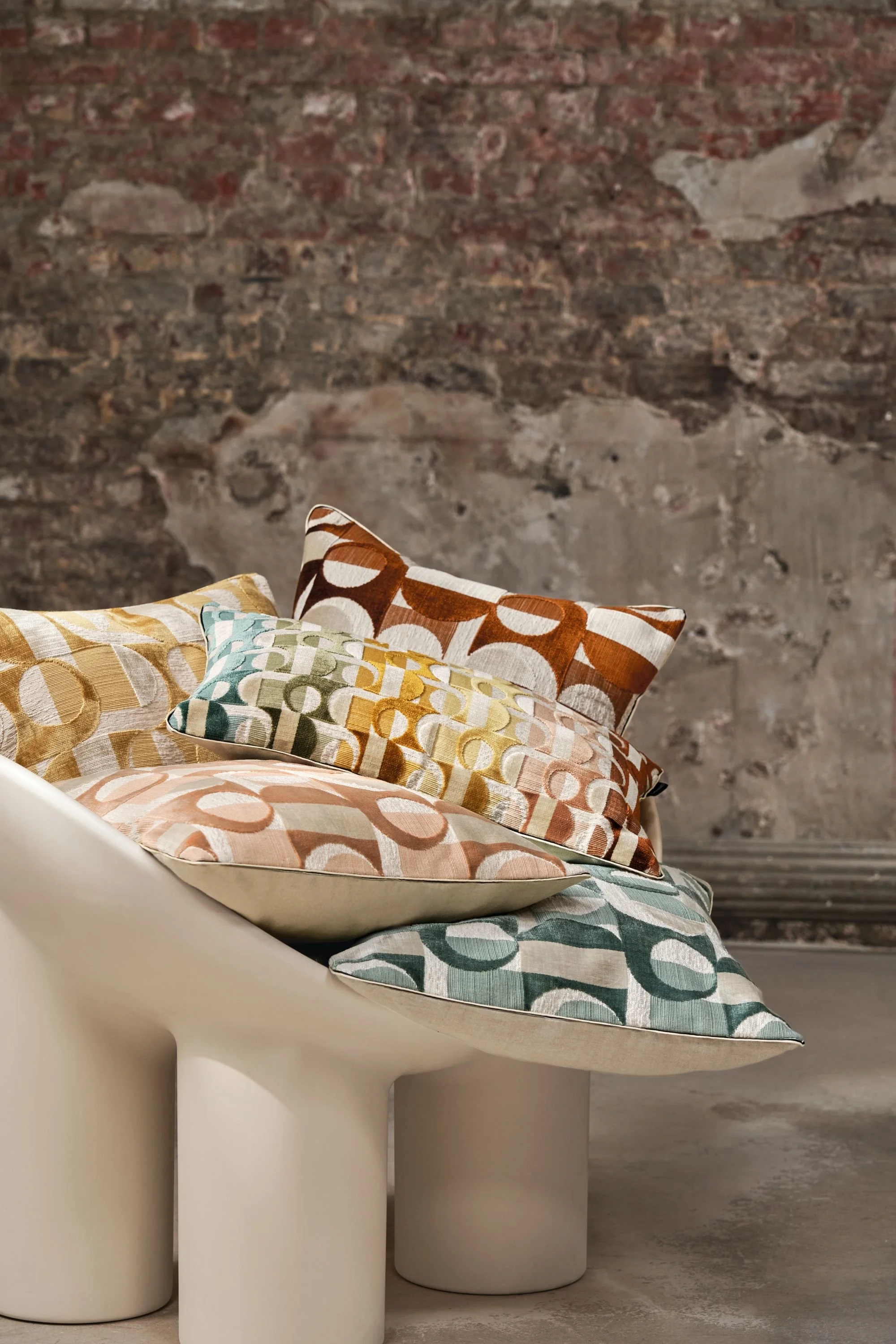 Maison Casamance Alliance embroidered cushions layered on a sculptural cream chair against a textured industrial backdrop.