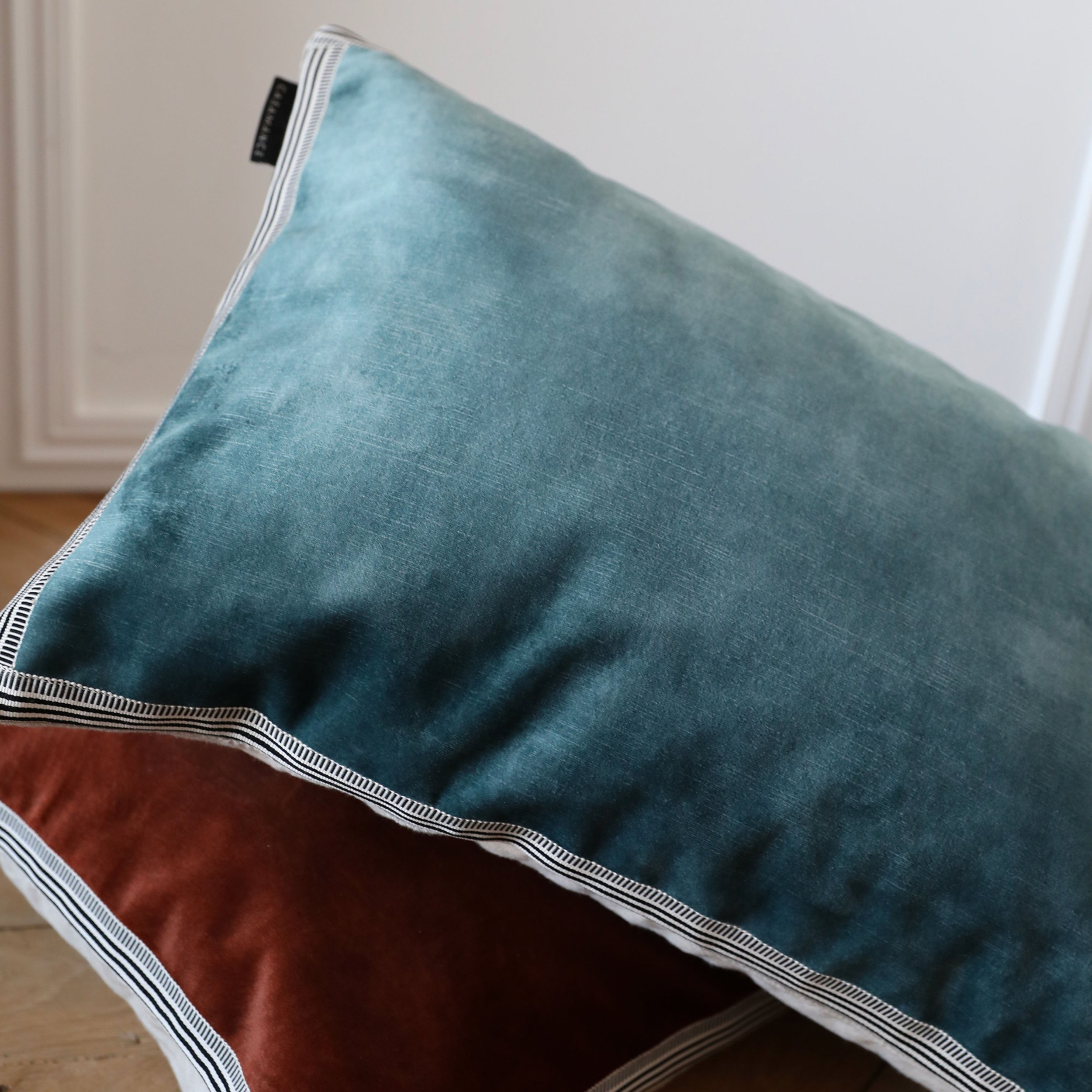 Close-up of a blue green Maison Casamance Manade velvet cushion resting on a copper one, showing the braid edge