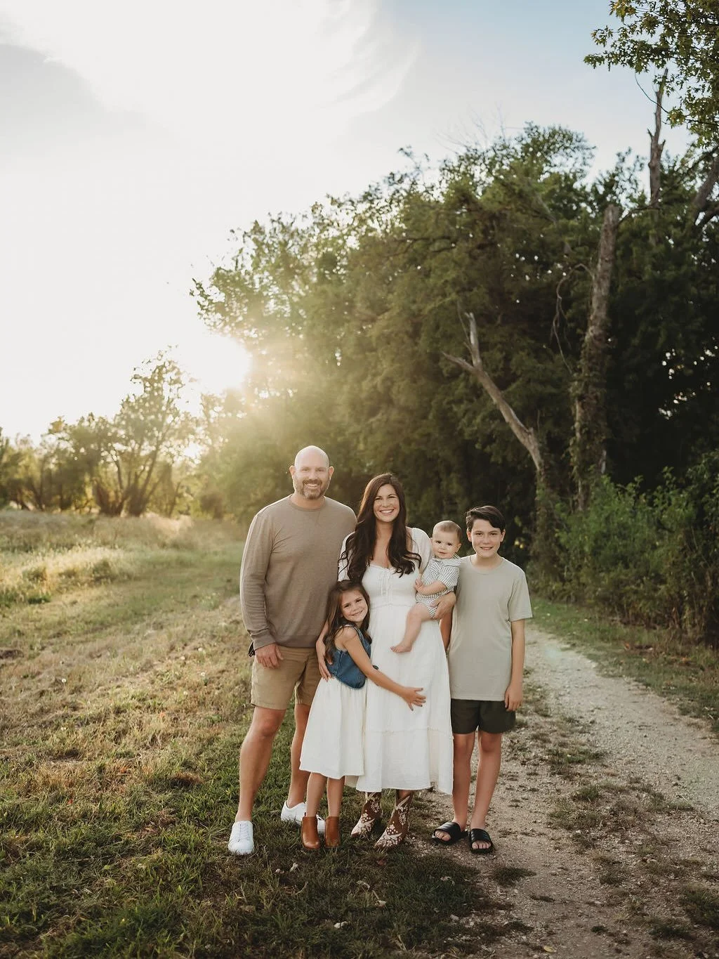A family of five standing outdoors on a dirt path near trees during sunset, smiling at the camera.