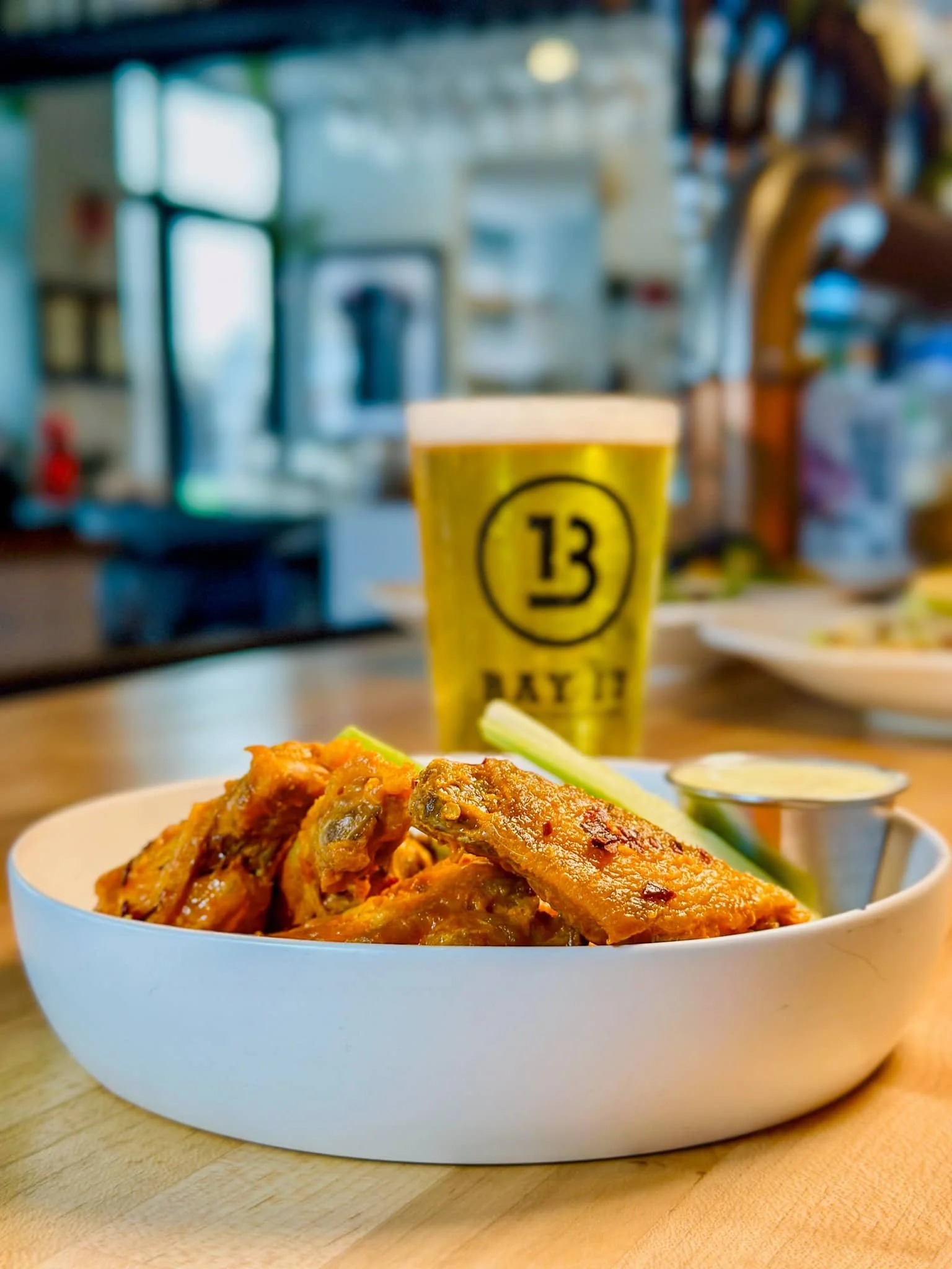 Close-up of an Asian dish, possibly chicken wings, served in a white bowl on a wooden table with a lime wedge and dipping sauce, a drink with the number 13 on a yellow cup in the background.