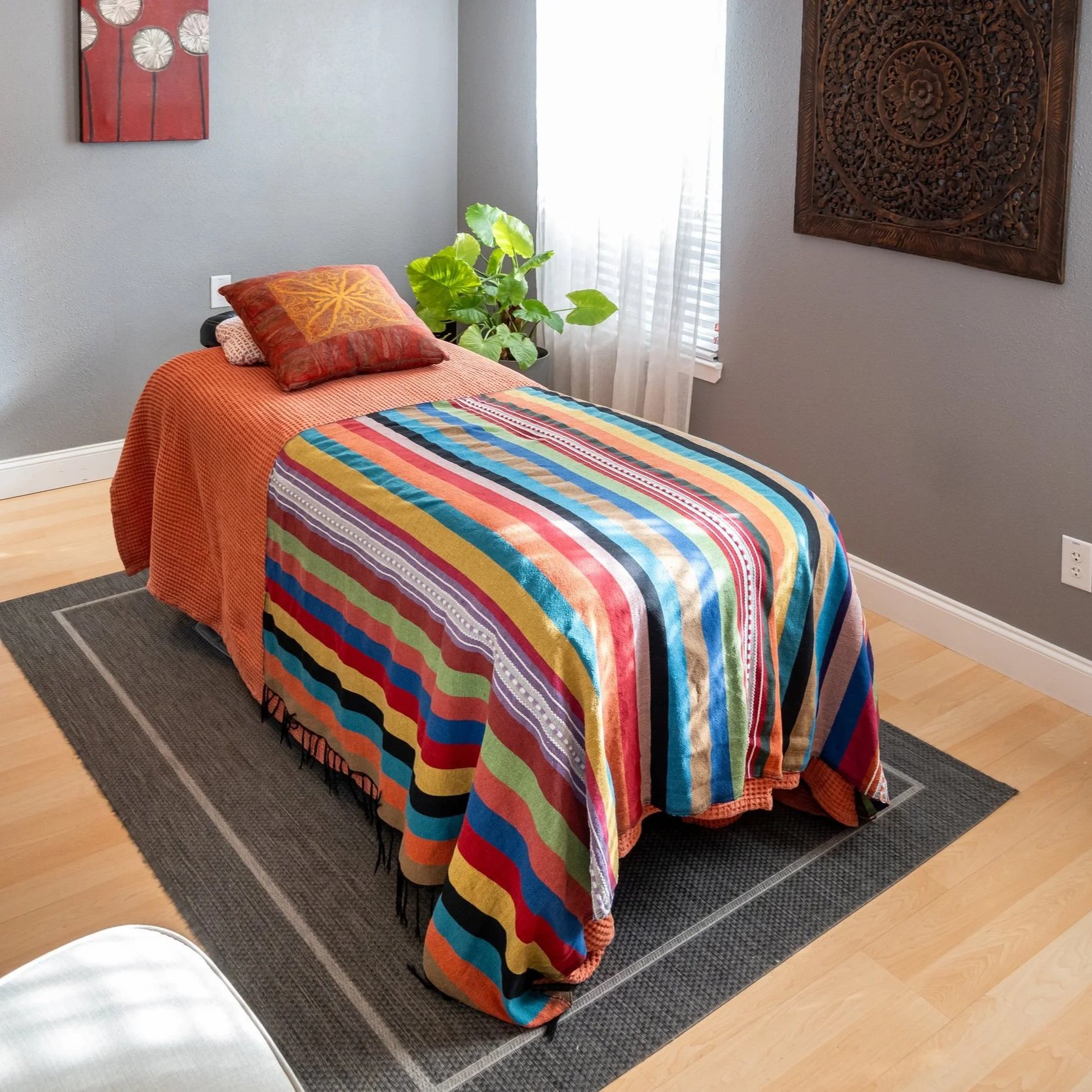A bedroom with a bed covered by a brightly striped quilt, paired with an orange bedspread and decorative pillows. There's a small black side table with a lamp, a plant near the window, and wall art on the walls. The room has light wood flooring and s