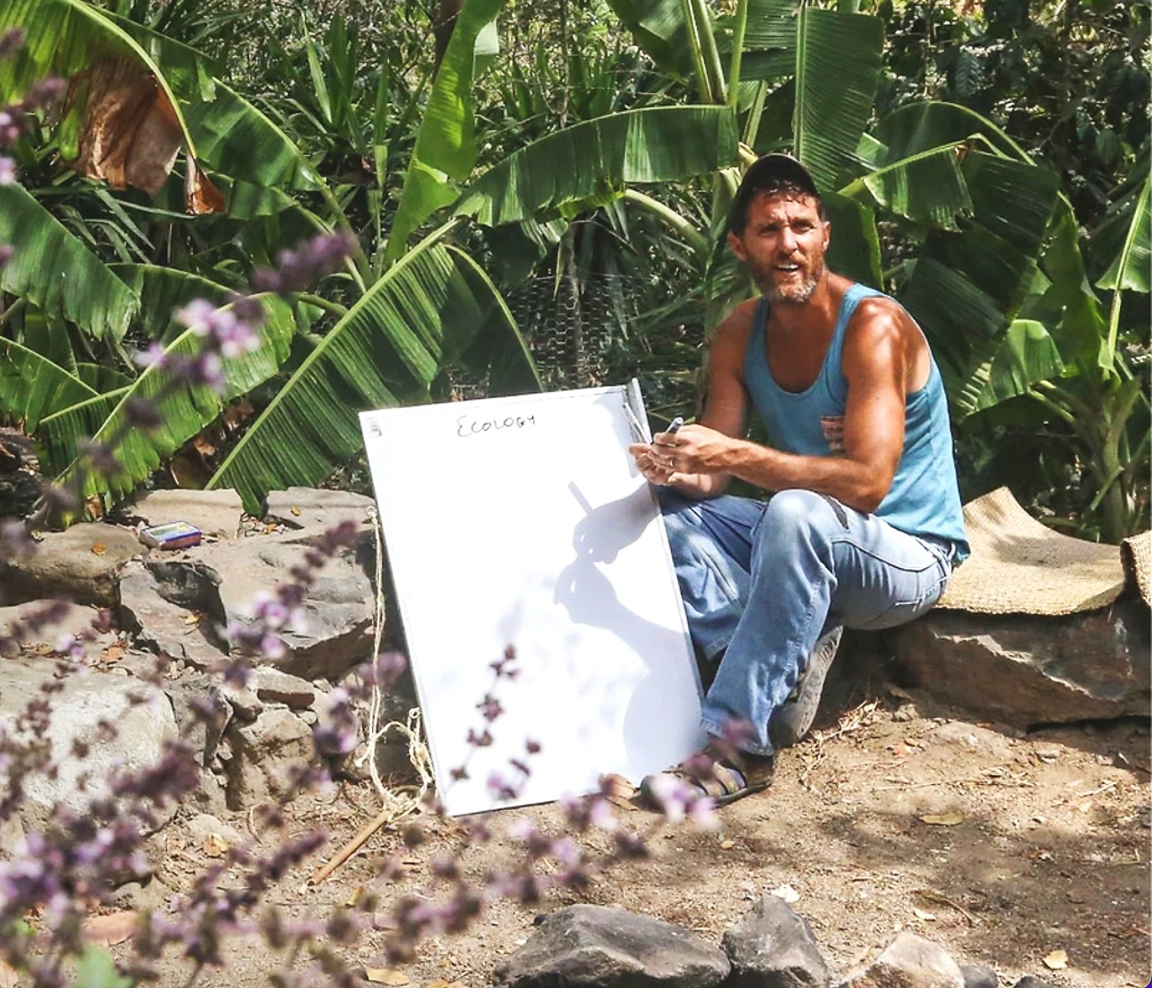 Neal, the permaculture farmer, sits down on a rock with a whiteboard in his hands on his farm in Guatemala.