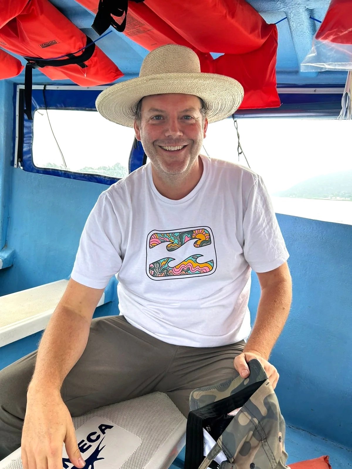 A man smiling and wearing a wide-brimmed straw hat, a white t-shirt with colorful abstract designs, and camouflage pants, sitting on a boat with red life jackets hanging above him and a blue interior, during an ecotour in Guatemala.