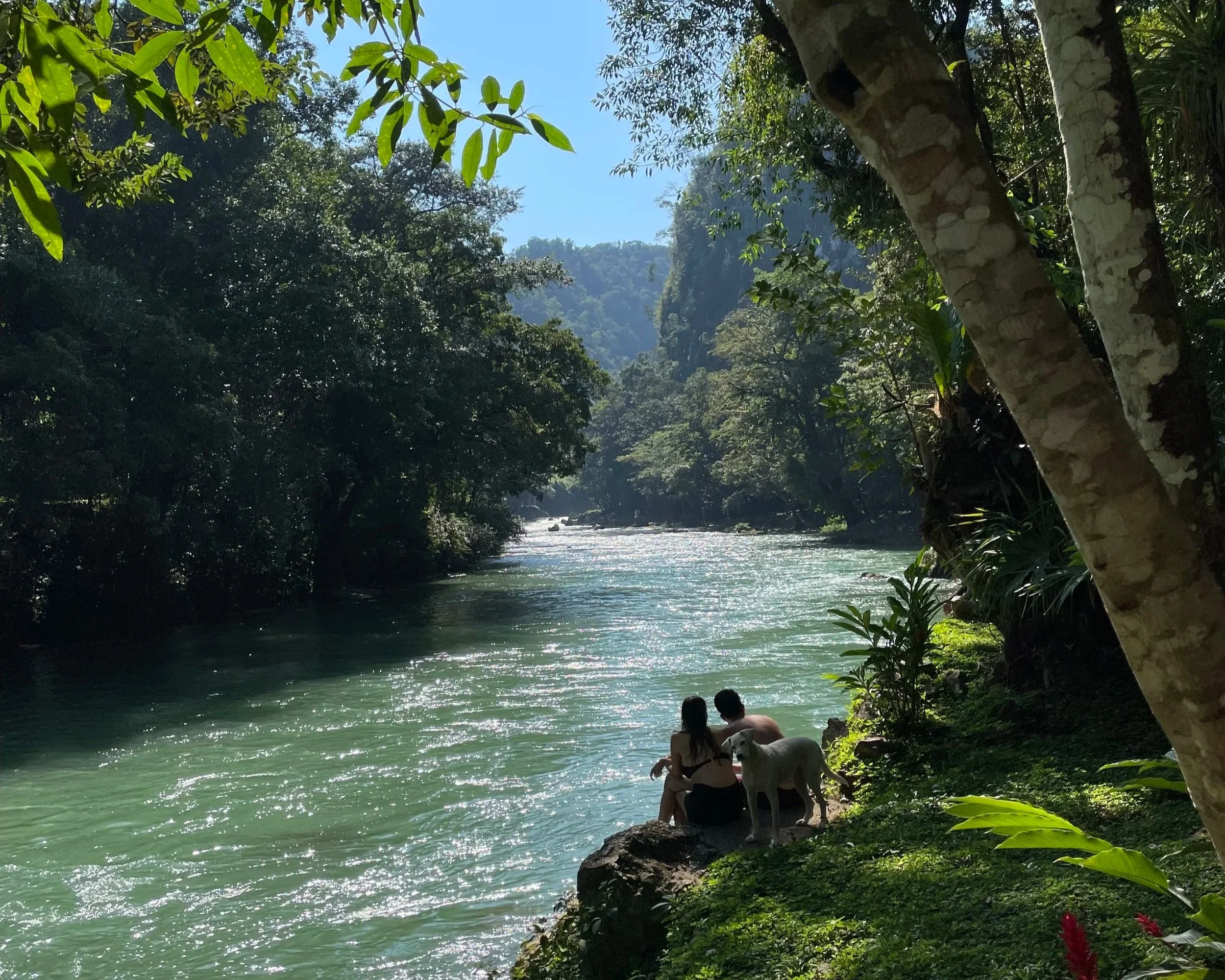 A couple is sitting on the river's edge with a dog looking at the camera in Semuc Champey