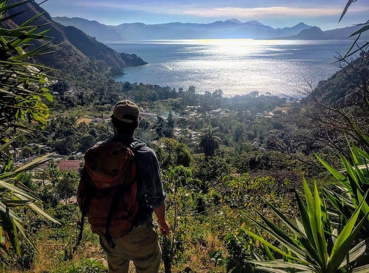 A man with a backpack is looking out toward the beautiful view of Lake Atitlan and surrounding mountains on a hike in the warm seasons of Guatemala