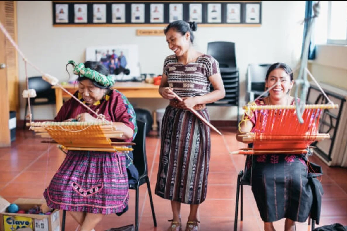 A woman stands over two Indigenous women as they are weaving on hand looms at Amigos de Santa Cruz, an organization you can donate to on Giving Tuesday