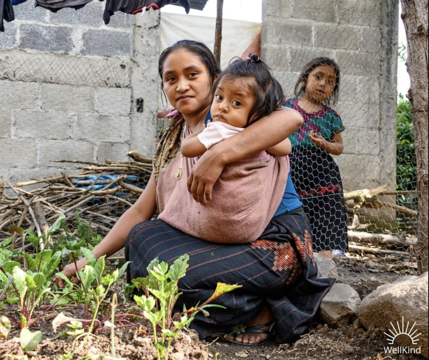 A mother is gardening in a hard while holding her baby in a pink sling, part of WellKind, an organization you can donate to on Giving Tuesday