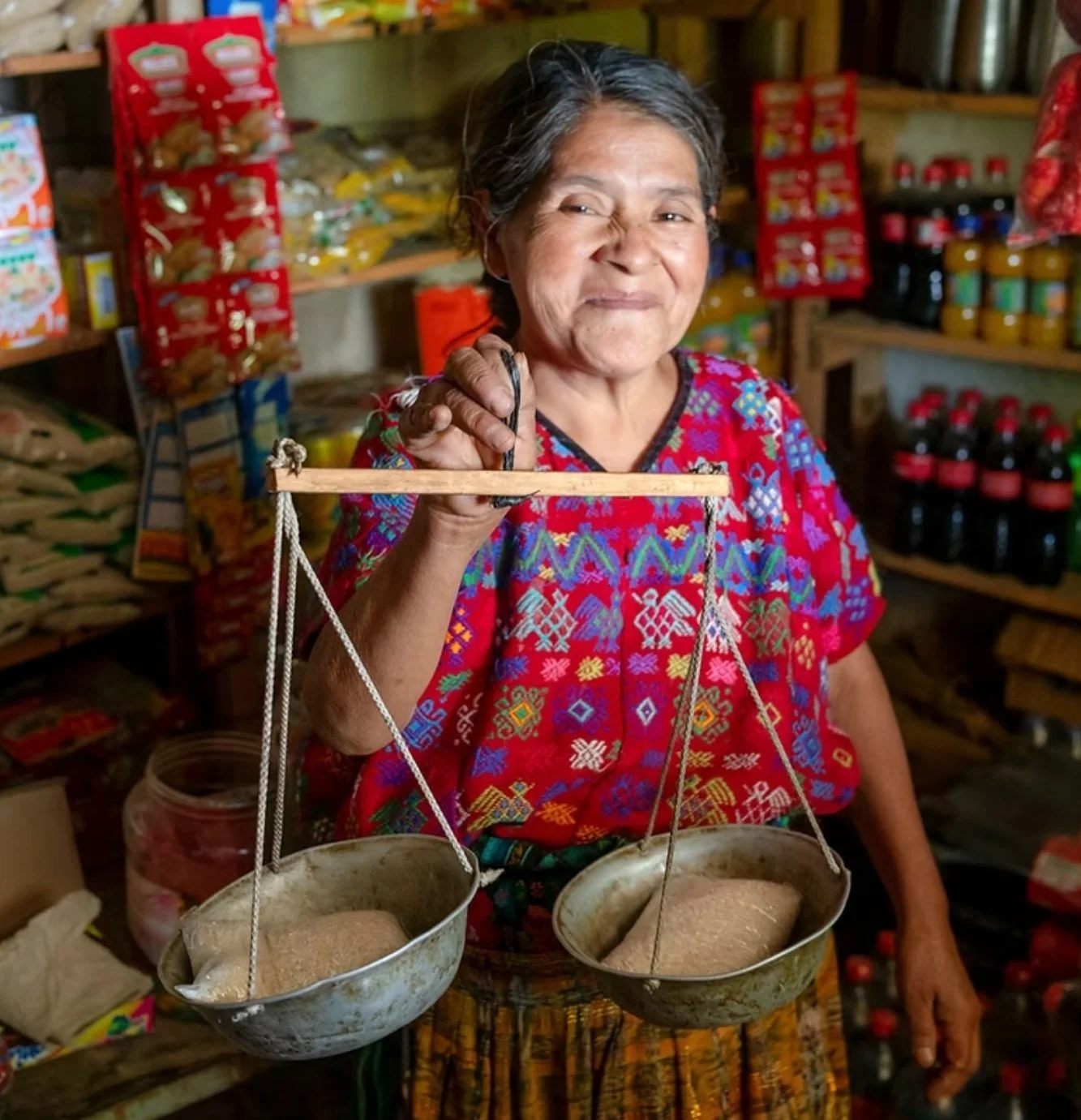 An Indigenious woman is in her shop holding a scale with products in each basket with WCCN, which you can donate to for Giving Tuesday