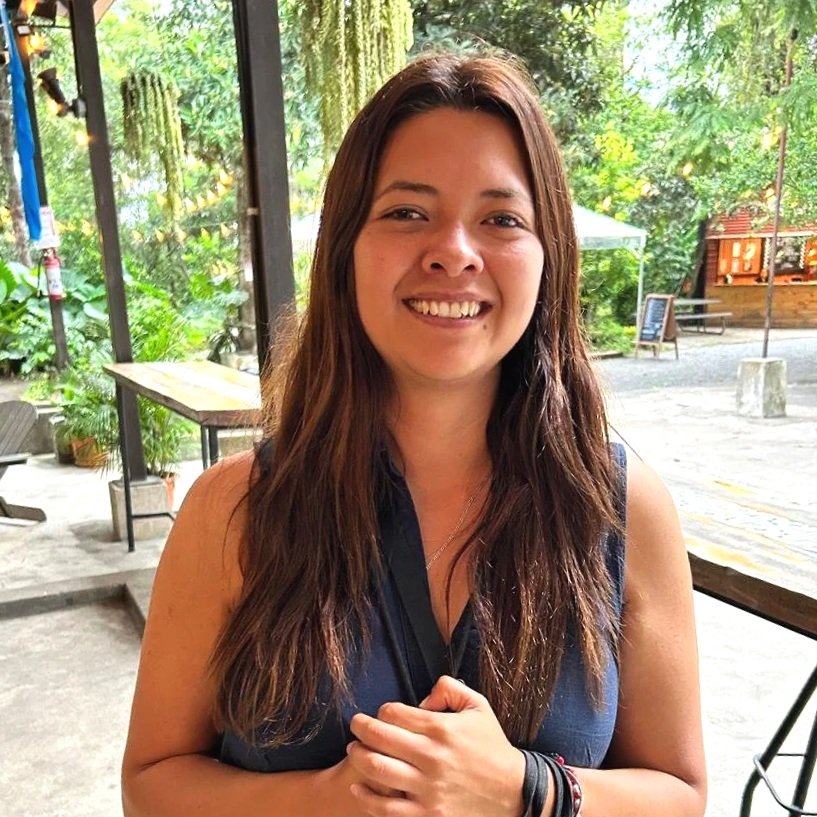 A woman with long brown hair smiling at the camera outdoors, surrounded by greenery and outdoor seating on a Naturally Smart Travel group tour in Guatemala.