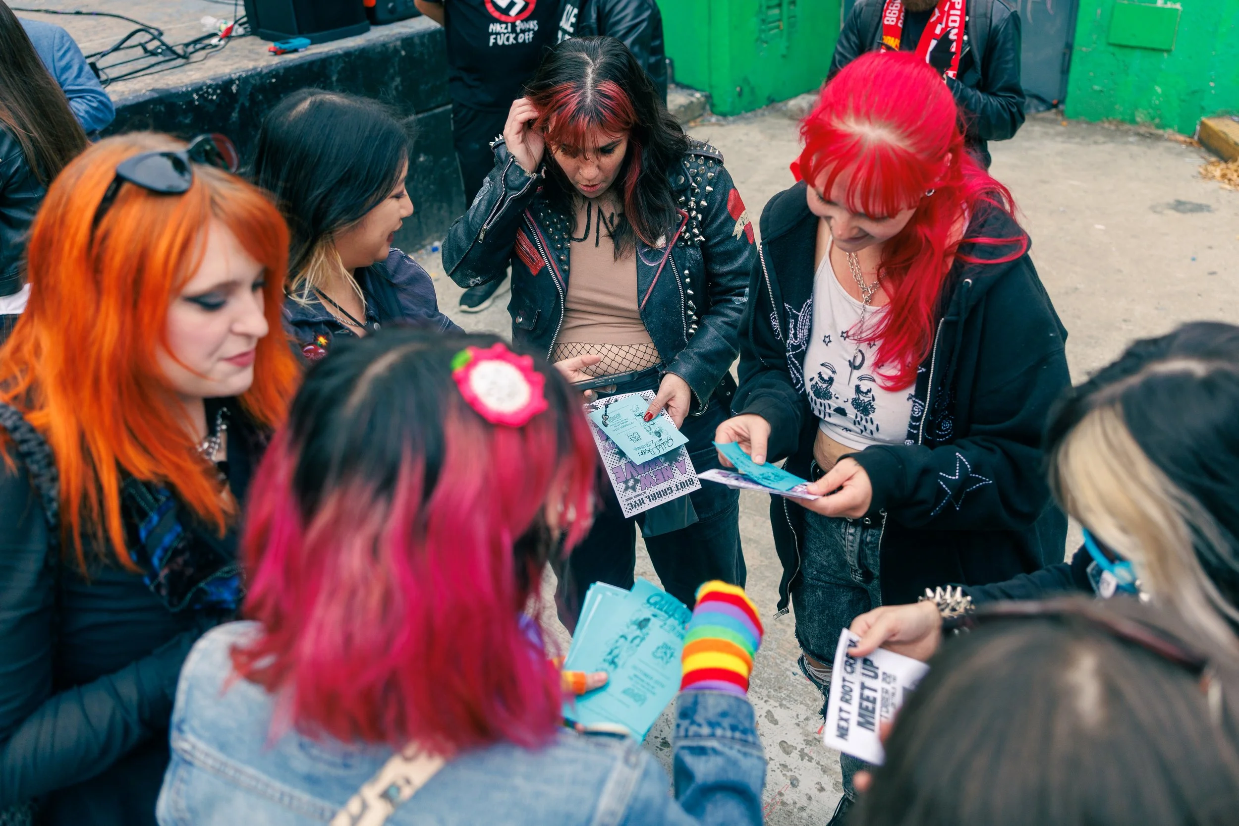 A group of women with colorful hair, some wearing leather jackets with spikes, gathered around looking at flyers or tickets, possibly at an outdoor event.