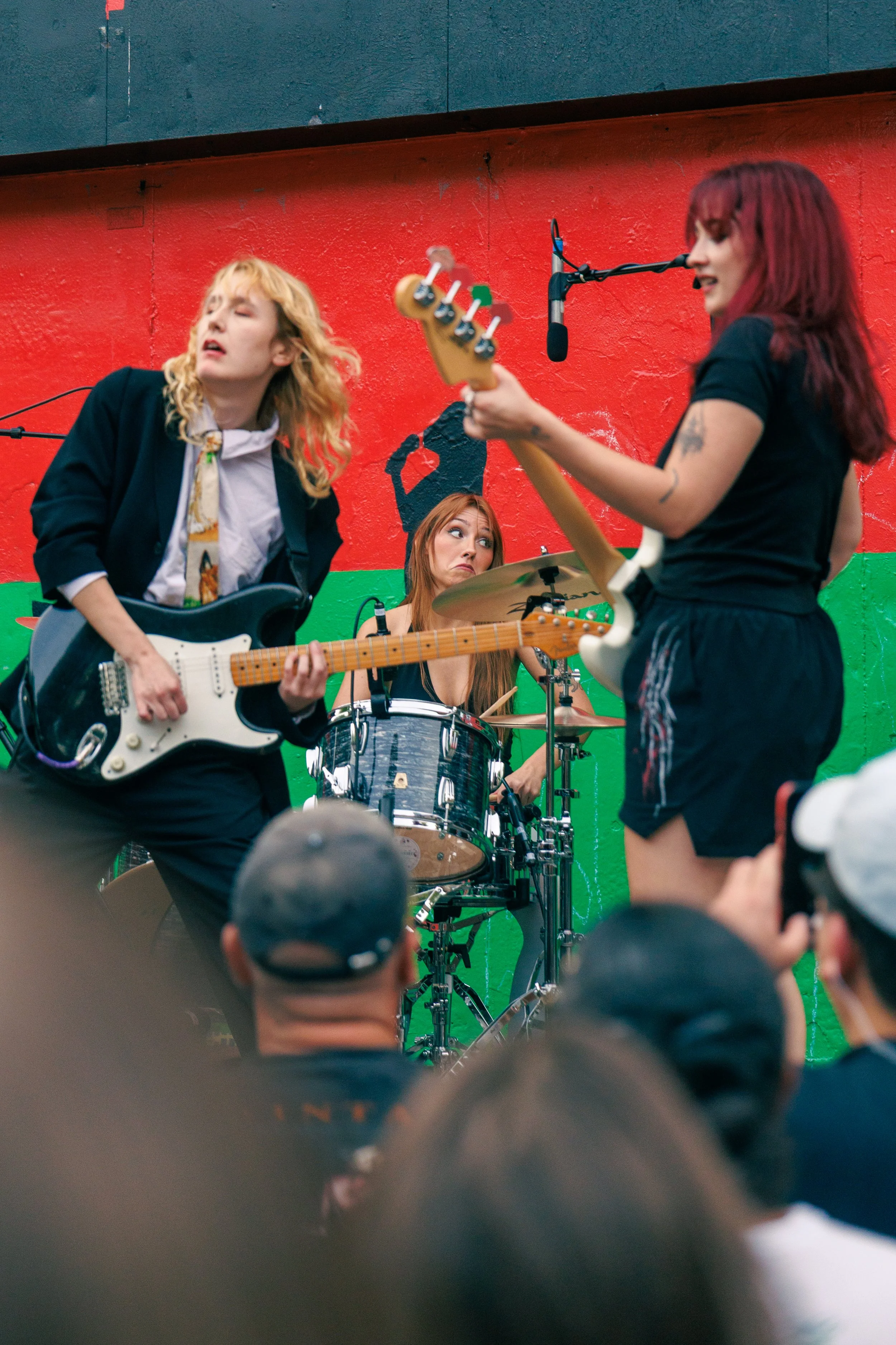 Three female musicians performing on stage with a red and green background, crowd in the foreground
