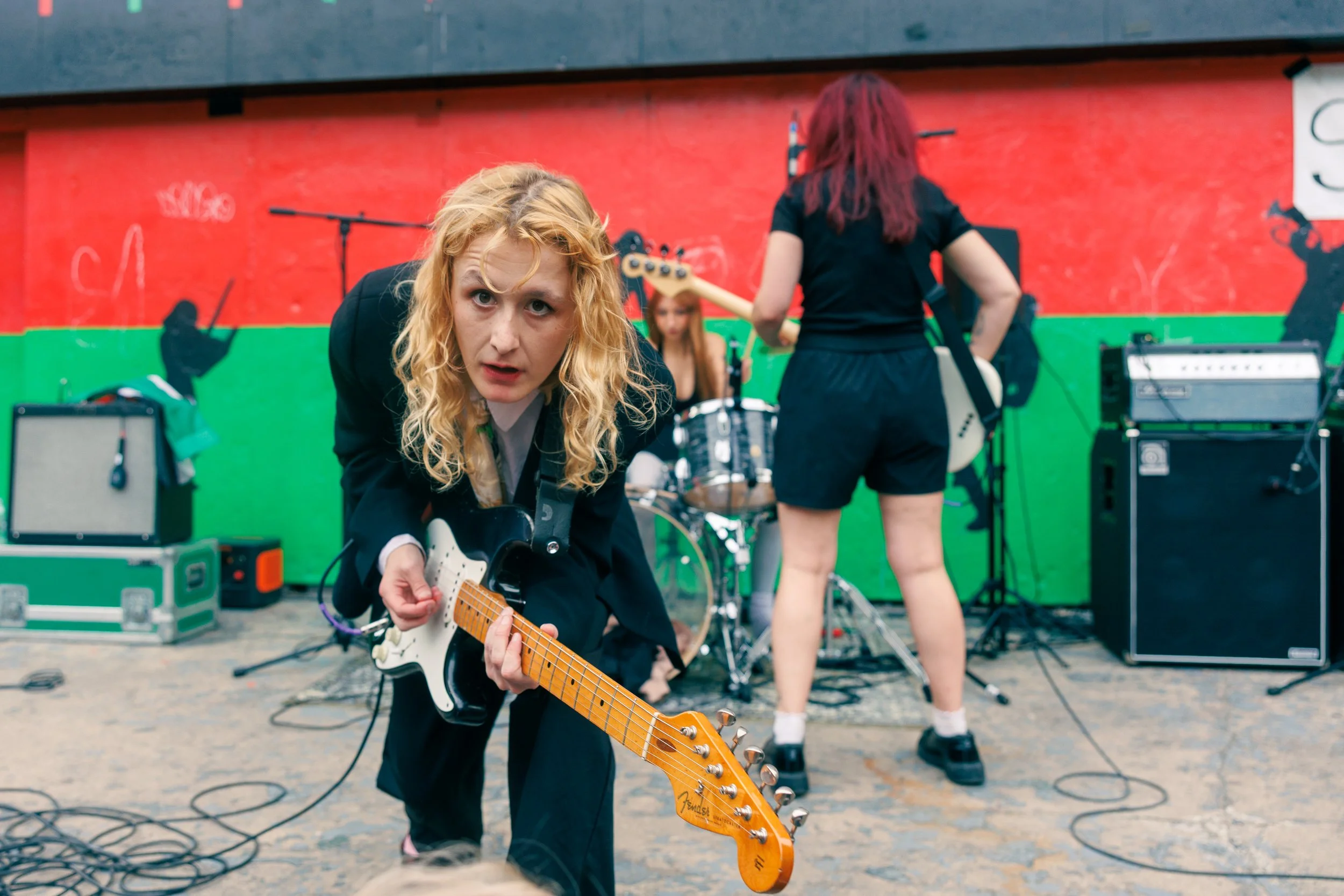 A woman with curly blonde hair playing an electric guitar in front of a band setup. The background features a red and green mural, and there are various musical equipment and instruments on stage.