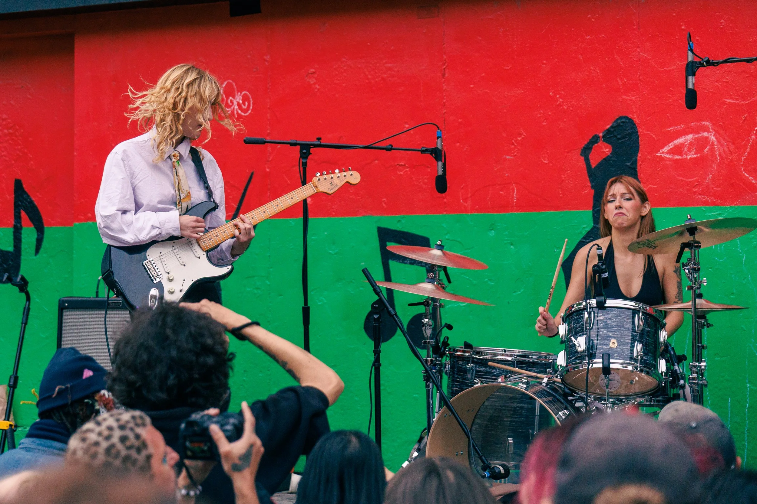 Two female musicians performing on a stage with a colorful wall background. The guitarist on the left is playing an electric guitar, and the drummer on the right is playing a drum set. There are microphones and audience members visible in the foregro