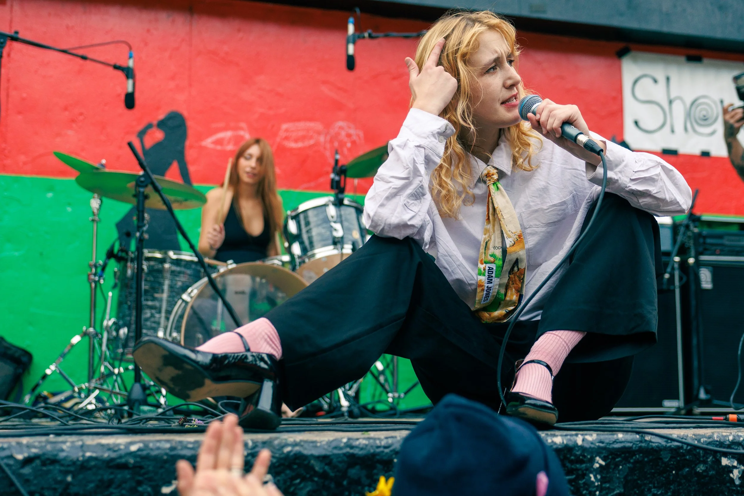 A young woman with curly blonde hair, wearing a white shirt, sitting on the floor with her legs crossed, speaking into a microphone during a performance. In the background, there is a colorful graffiti wall and a drummer woman with long red hair play