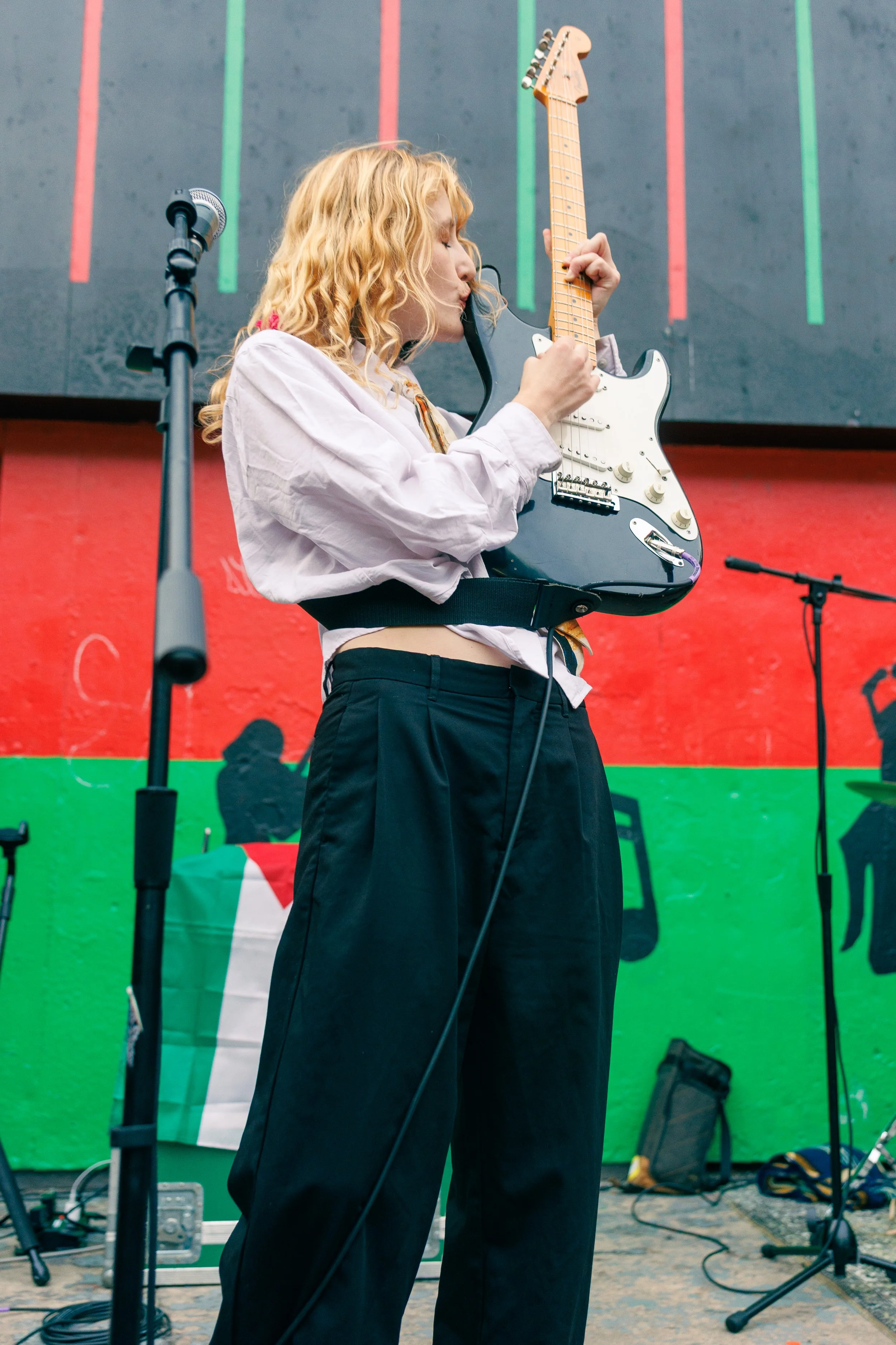 A woman with long blonde curly hair, wearing a white shirt and black pants, is playing an electric guitar on stage. There is a microphone stand in front of her, and a green and red mural with an Italian flag motif in the background.