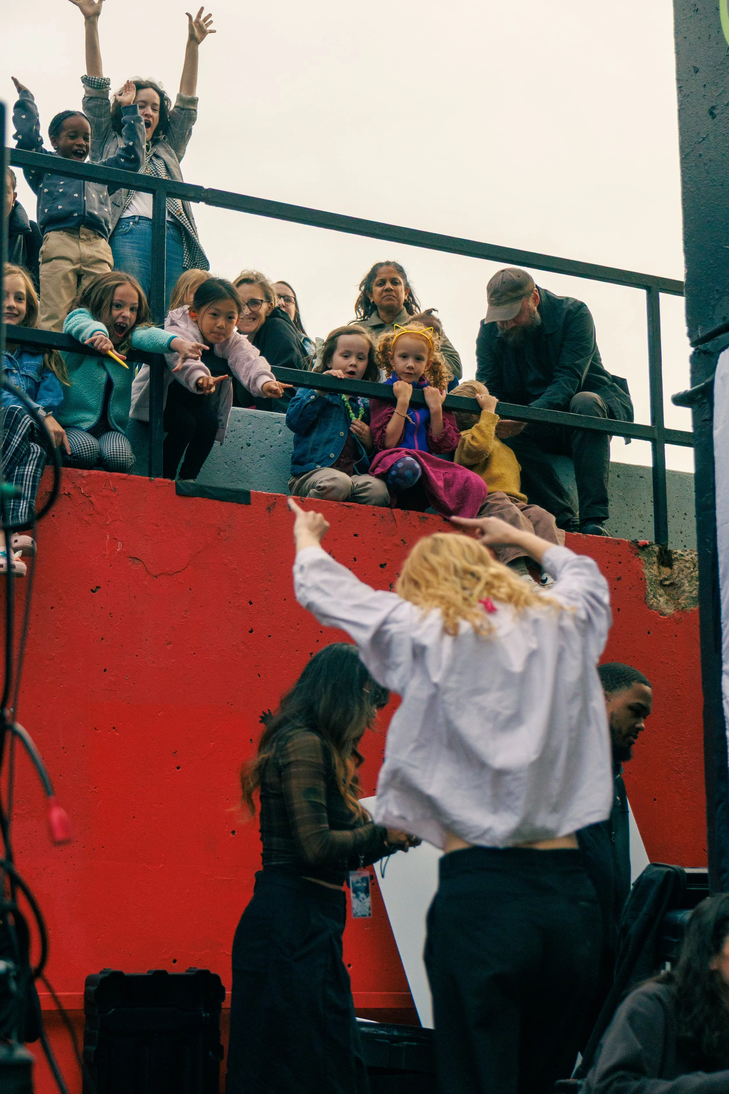 Group of children and adults on a balcony, reaching down towards someone below. The children appear excited and animated, with some pointing and others reaching out. The adults are observing or engaging with the children.