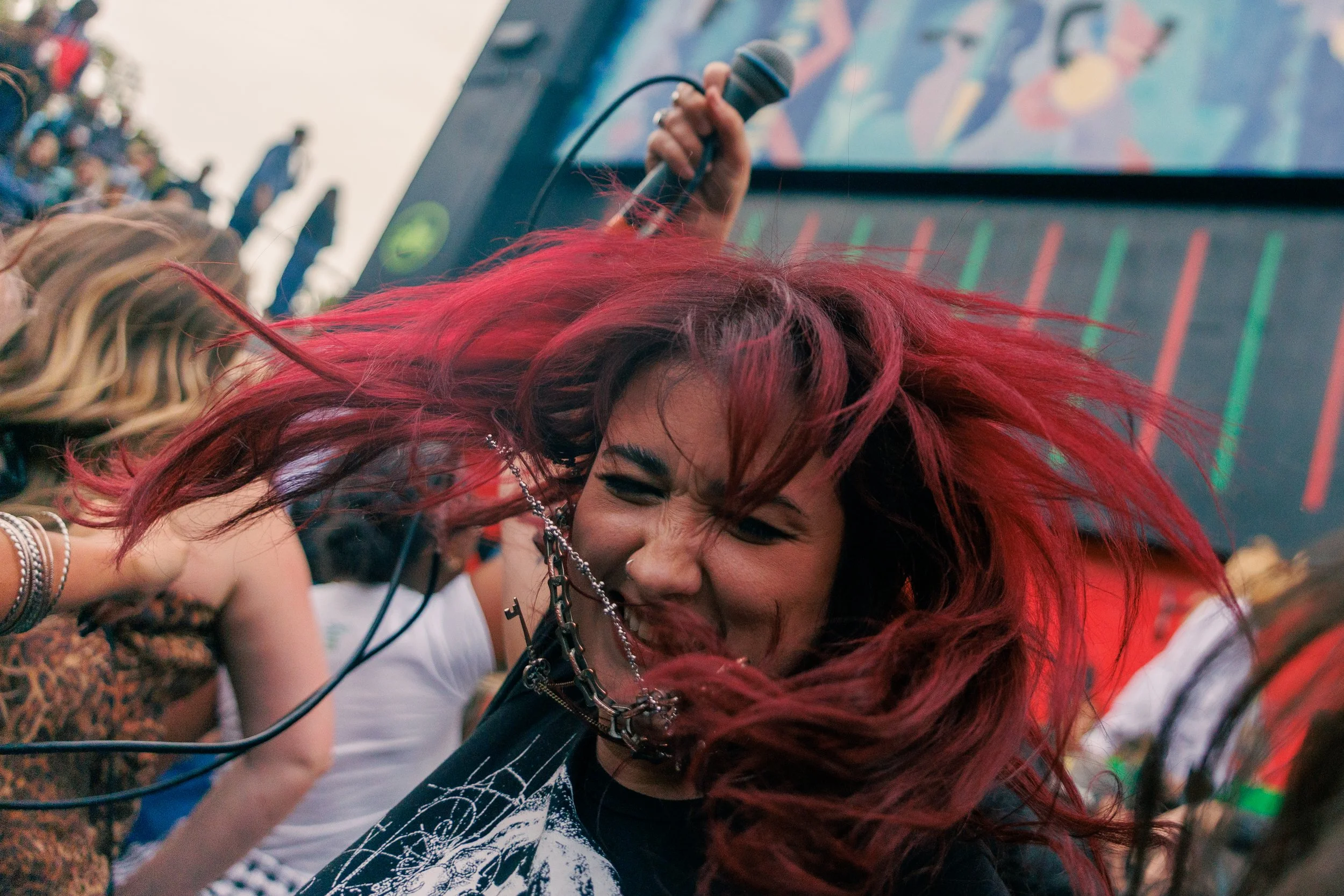 A woman with vibrant red hair and gothic style jewelry, smiling as she leans forward at an outdoor event with a crowd in the background. Part of her face is visible as she appears to be laughing or singing.