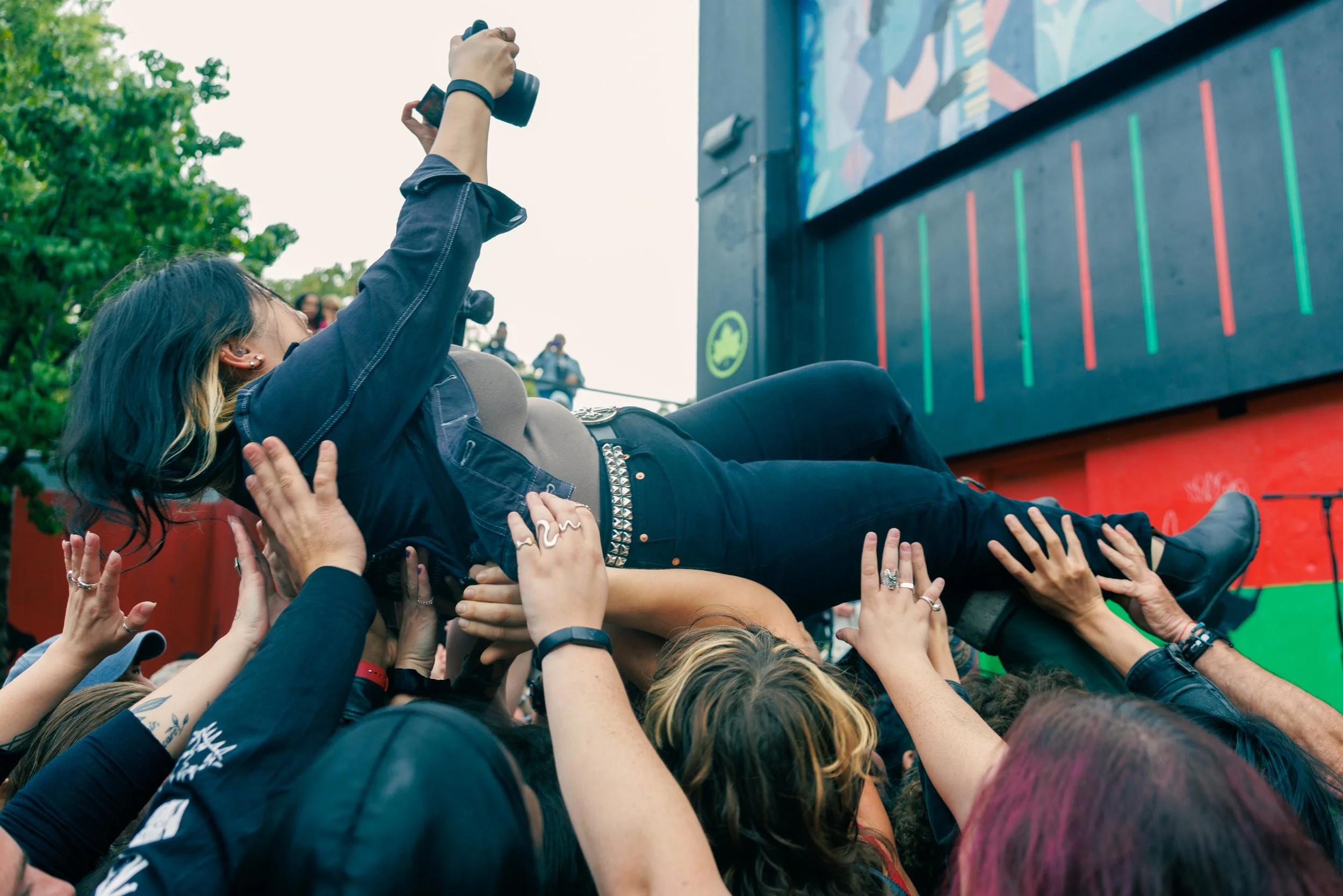 Person lying on top of a crowd during a music festival, reaching out with one hand, surrounded by fans' hands, outdoors with trees in the background.