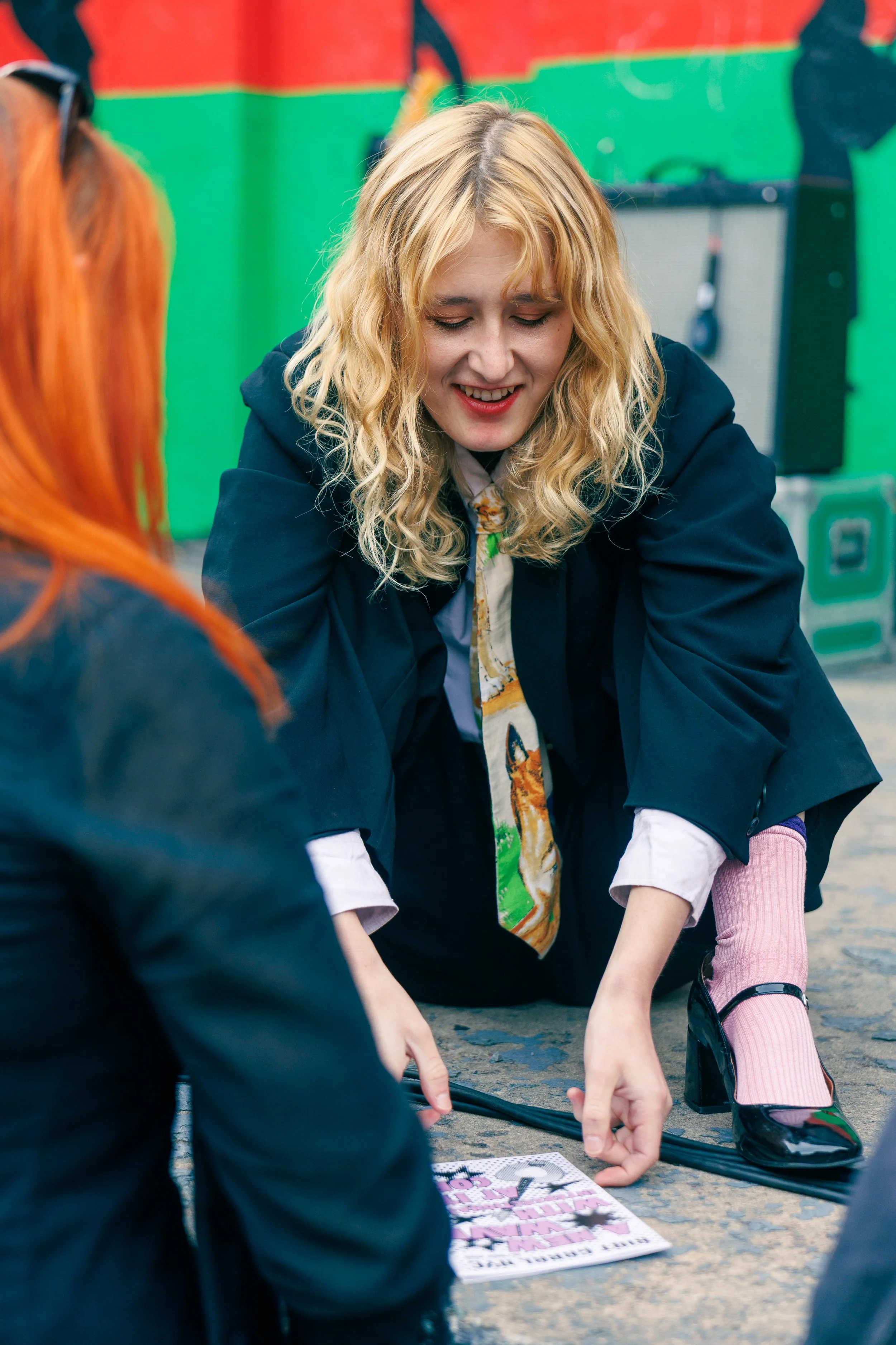 A woman with blonde curly hair and a black coat is squatting down and smiling while interacting with a small child. The child is wearing pink tights and black shoes. They are on a paved surface with a colorful background, and there are some books on 