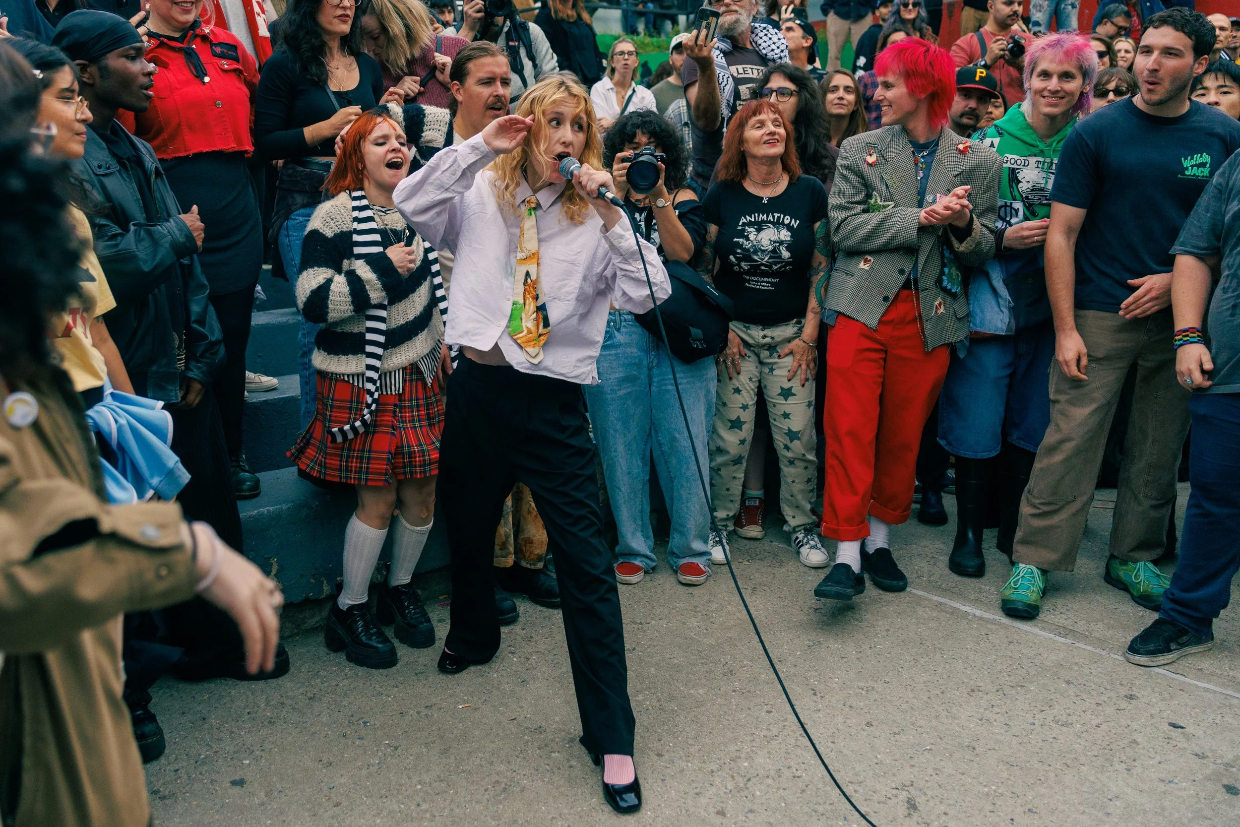 A crowd of people gathered outdoors, with some holding cameras and microphones. One woman with blonde hair is speaking into a microphone while others watch and listen. The group appears engaged in a public event or protest.