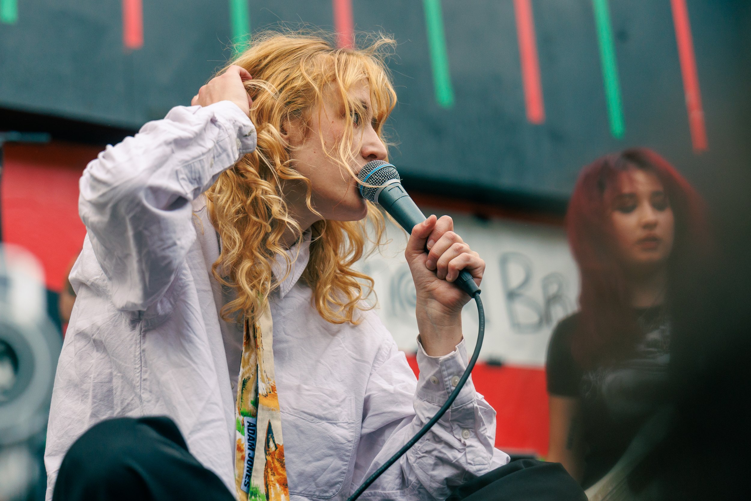 A woman with curly red hair wearing a white shirt and a colorful scarf, sitting on the ground, holding a microphone close to her mouth, possibly speaking or singing at an event with a background of laundry machines and neon lights.