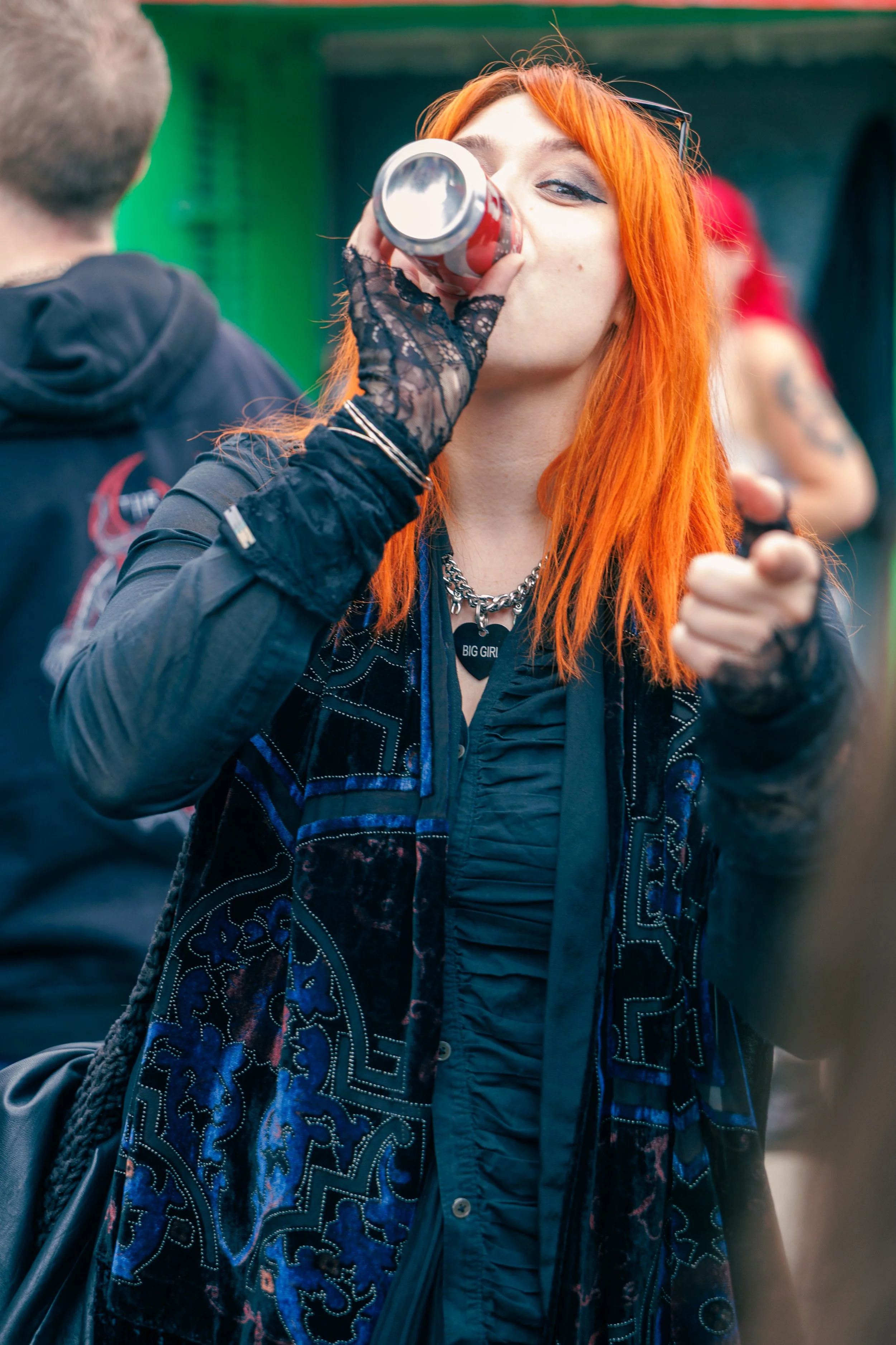 A person with orange hair wearing a black jacket and gloves, drinking a beverage from a can, with a person in a red shirt blurred in the background.