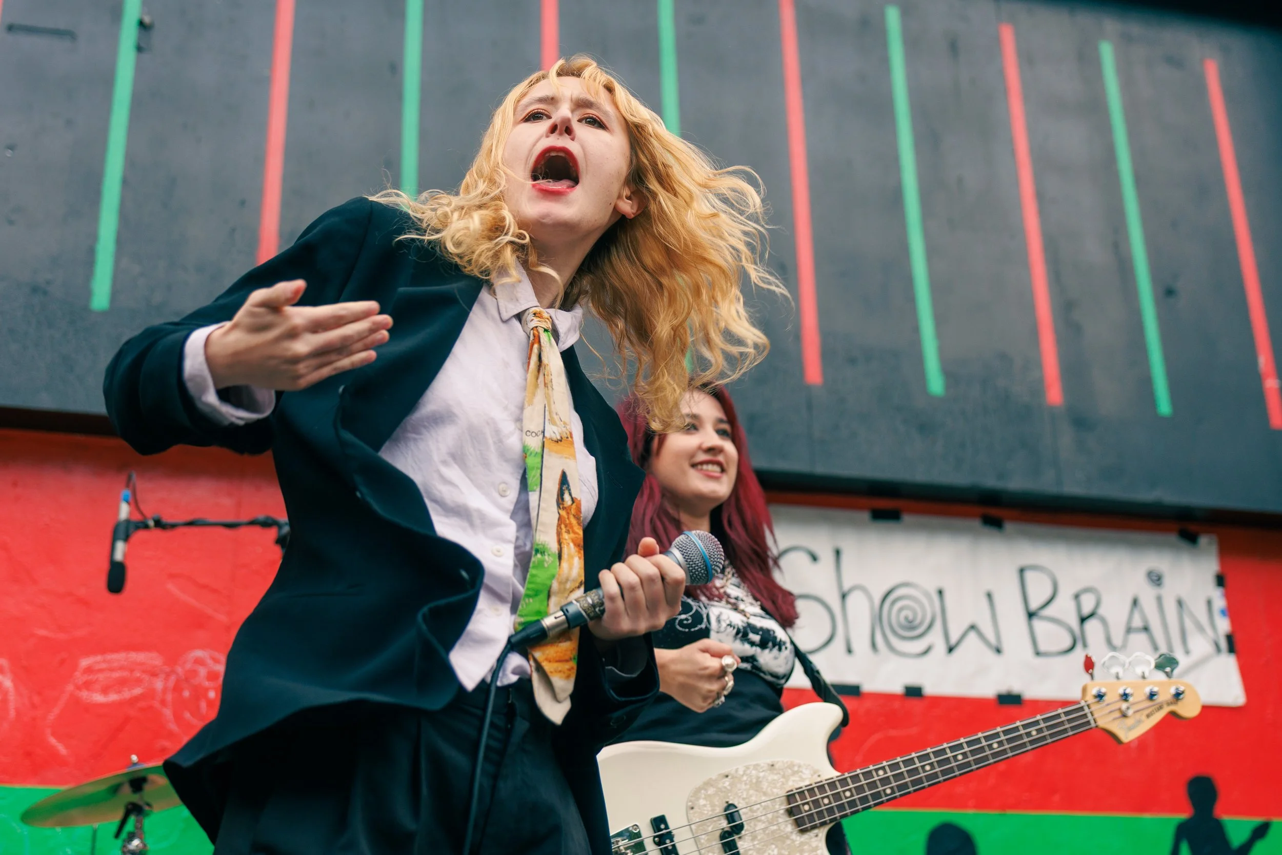 Two women participating in a protest or rally, one is speaking passionately into a microphone, and the other is holding a guitar. They are standing in front of a decorated board with red and green lines and writing, possibly at a public demonstration