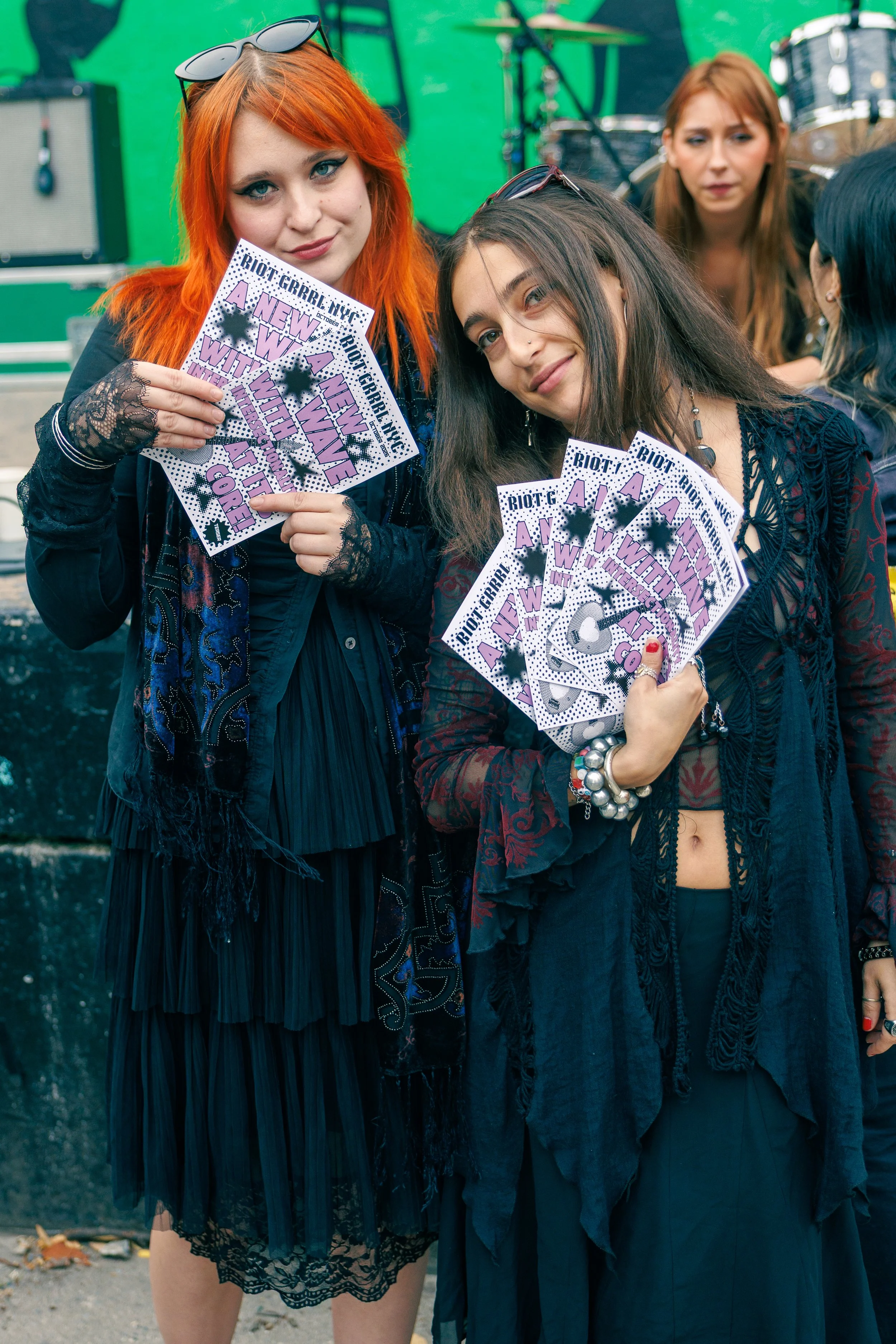 Three young women standing outdoors at a music event, holding purple and white protest signs with slogans. The woman in the foreground has long brown hair, a nose piercing, and wears a dark, lace-trimmed skirt and a dark vest. The woman to her left h