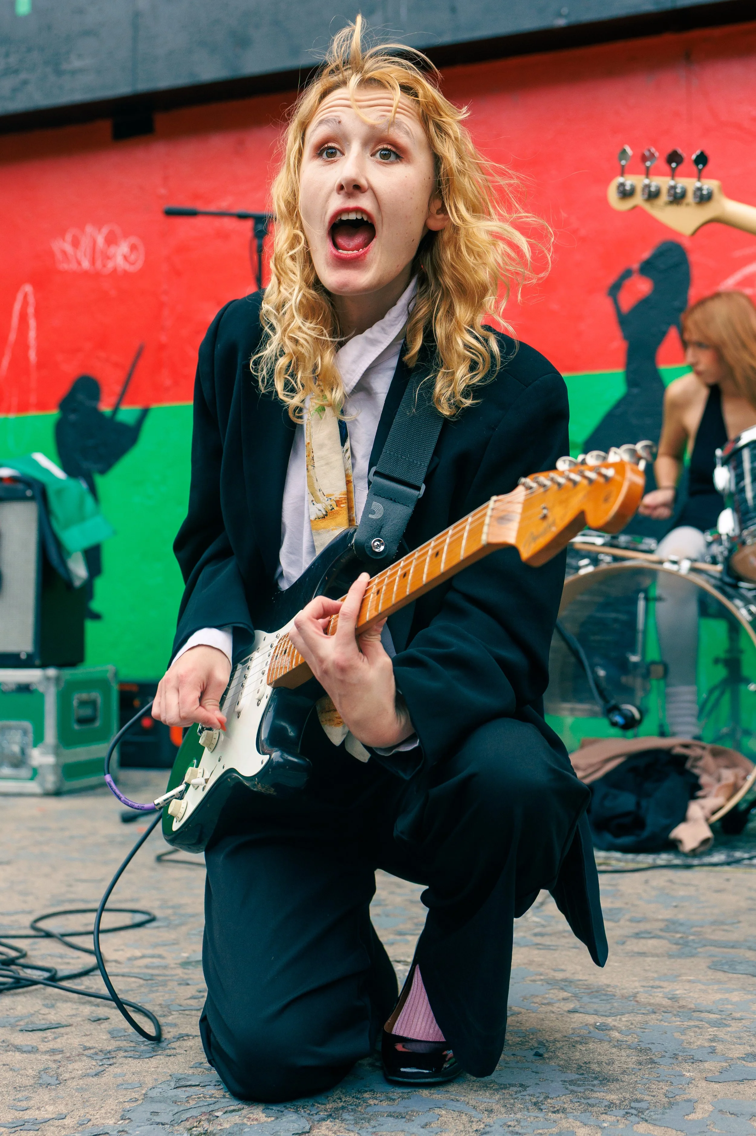 A woman performing live music outdoors, playing an electric guitar, with a woman in the background near musical equipment, and a colorful mural on the wall.