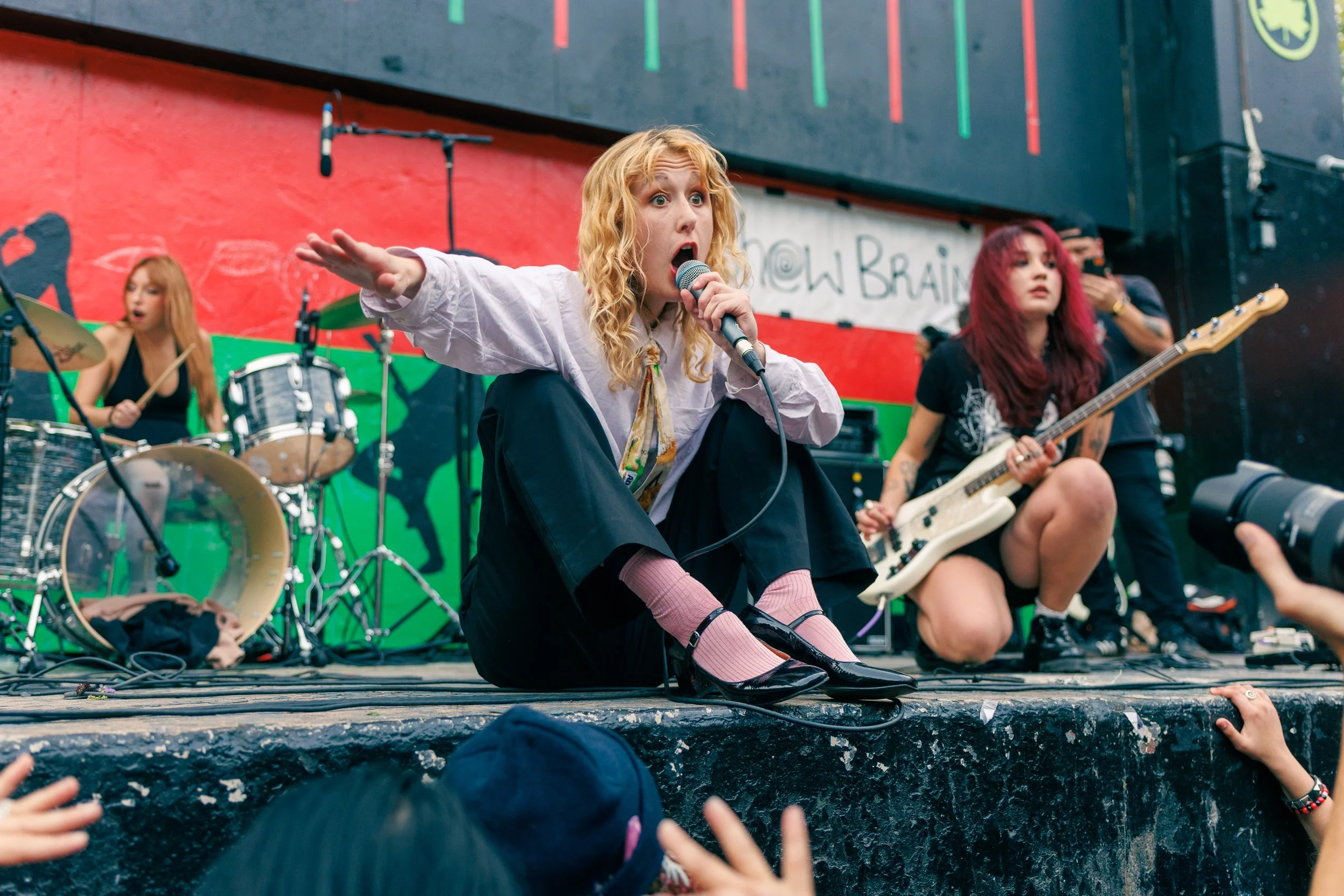 A young woman with curly blonde hair and pink socks is sitting on the floor with a microphone in her hand, singing or speaking to an audience. She is surrounded by a band playing musical instruments on a stage with a colorful background and attendees