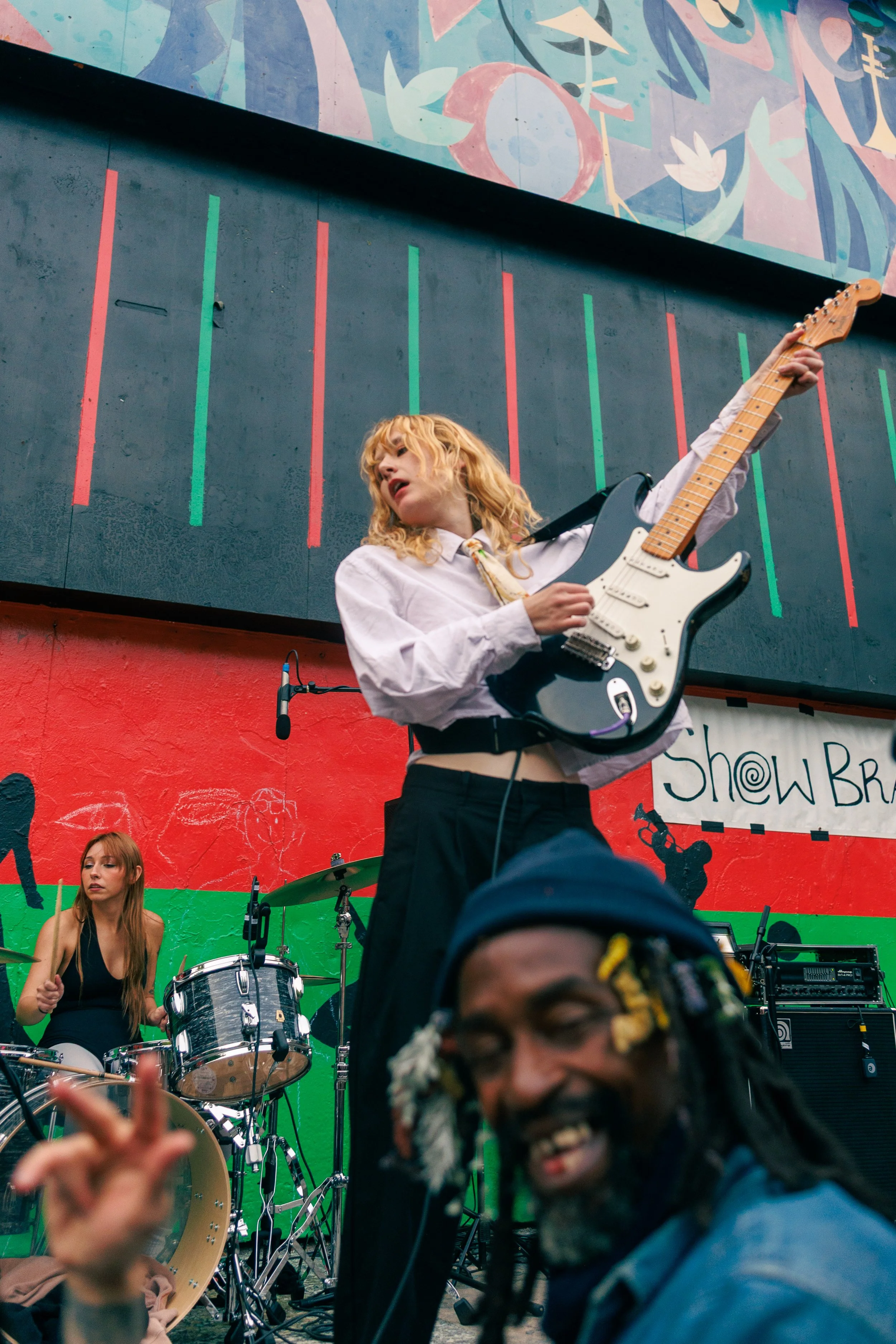 A woman with curly blonde hair playing an electric guitar on stage during a live performance, with a vibrant colorful mural in the background, and a woman playing drums visible behind her.