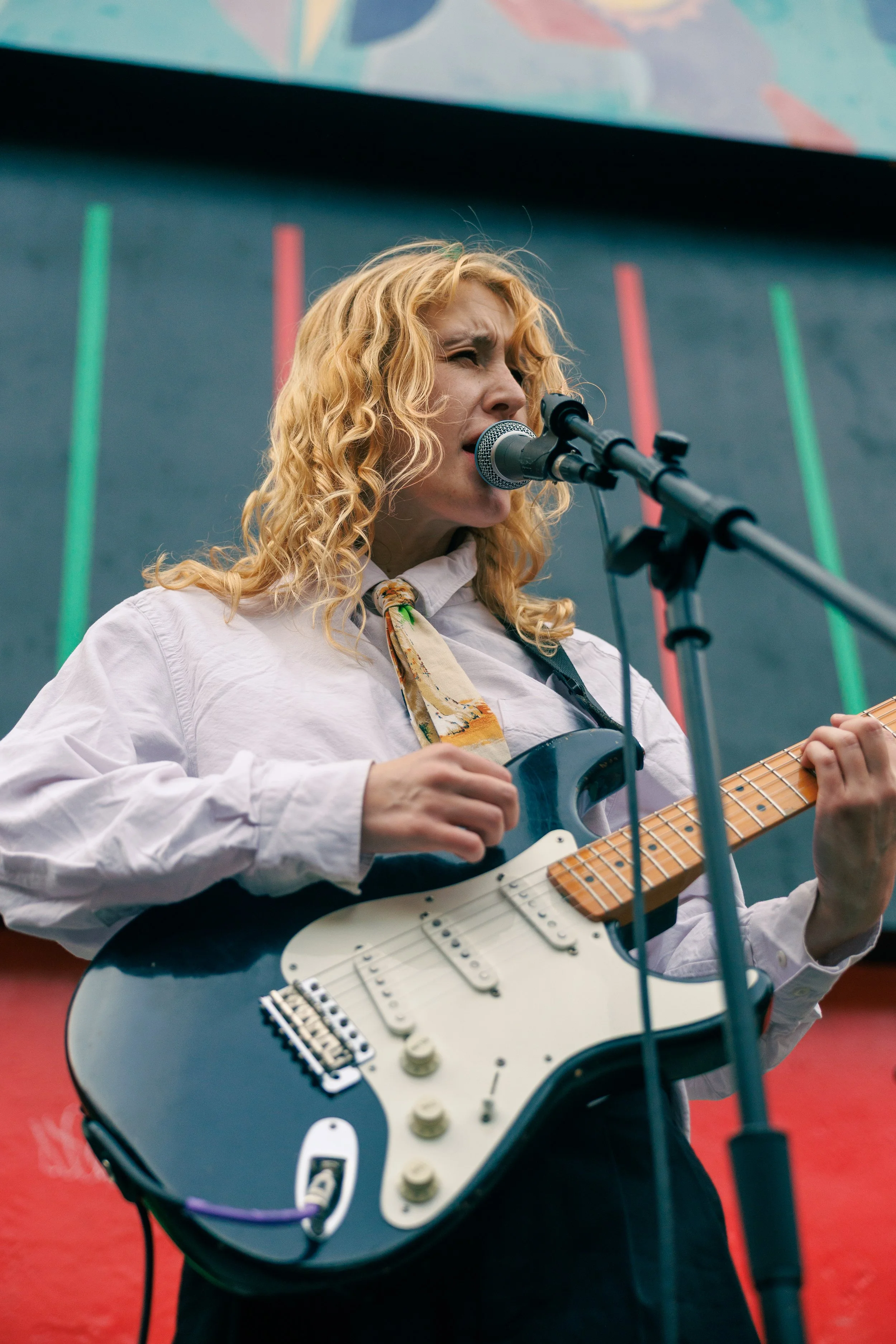 A woman with curly blonde hair playing an electric guitar and singing into a microphone at a live performance. She is wearing a white shirt and a patterned scarf, with a backdrop featuring colorful vertical lines.