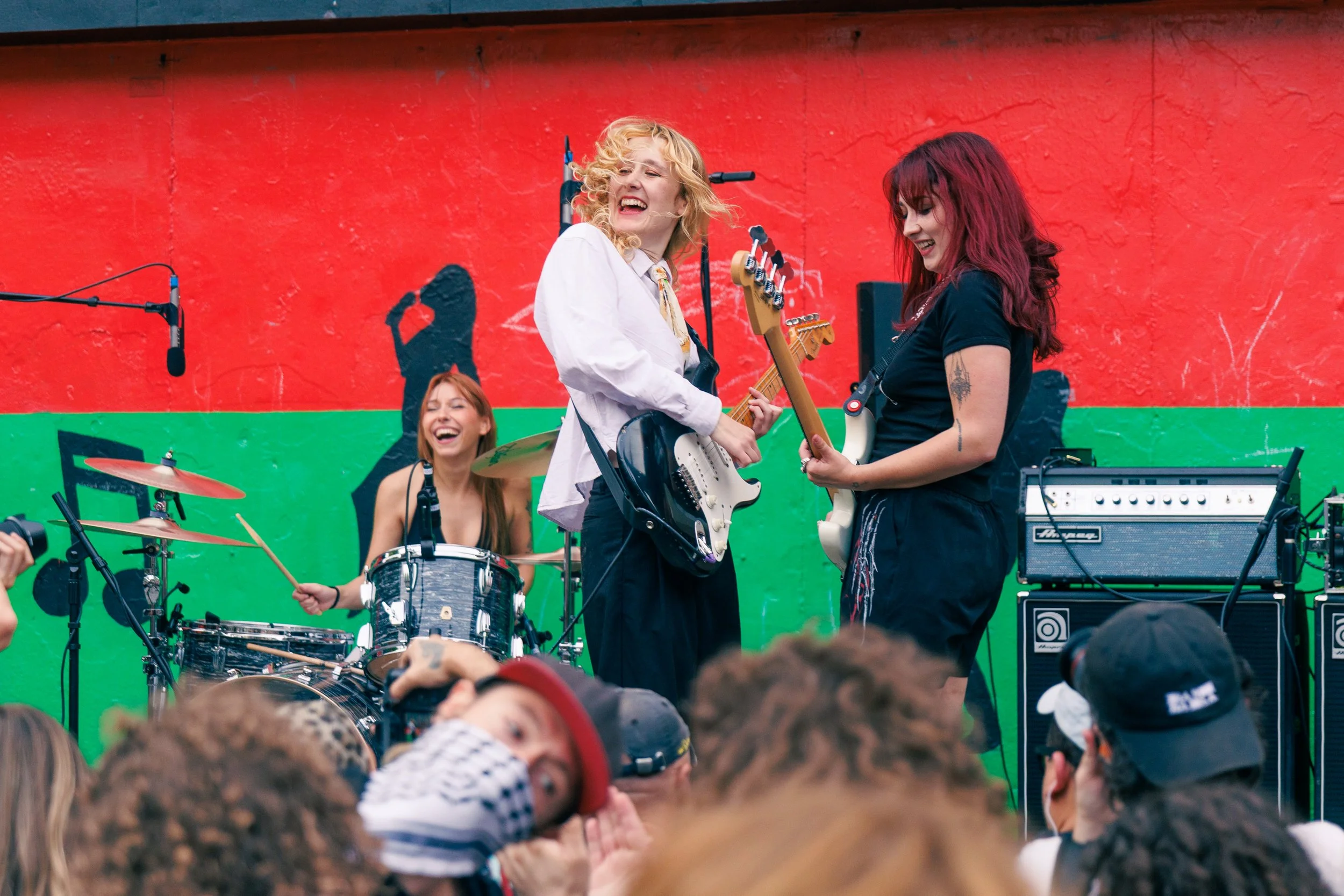 Three women perform on stage with musical instruments in front of a red and green painted wall. One woman is playing drums, another is playing an electric guitar, and the third is playing a bass guitar. The audience is visible in the foreground.