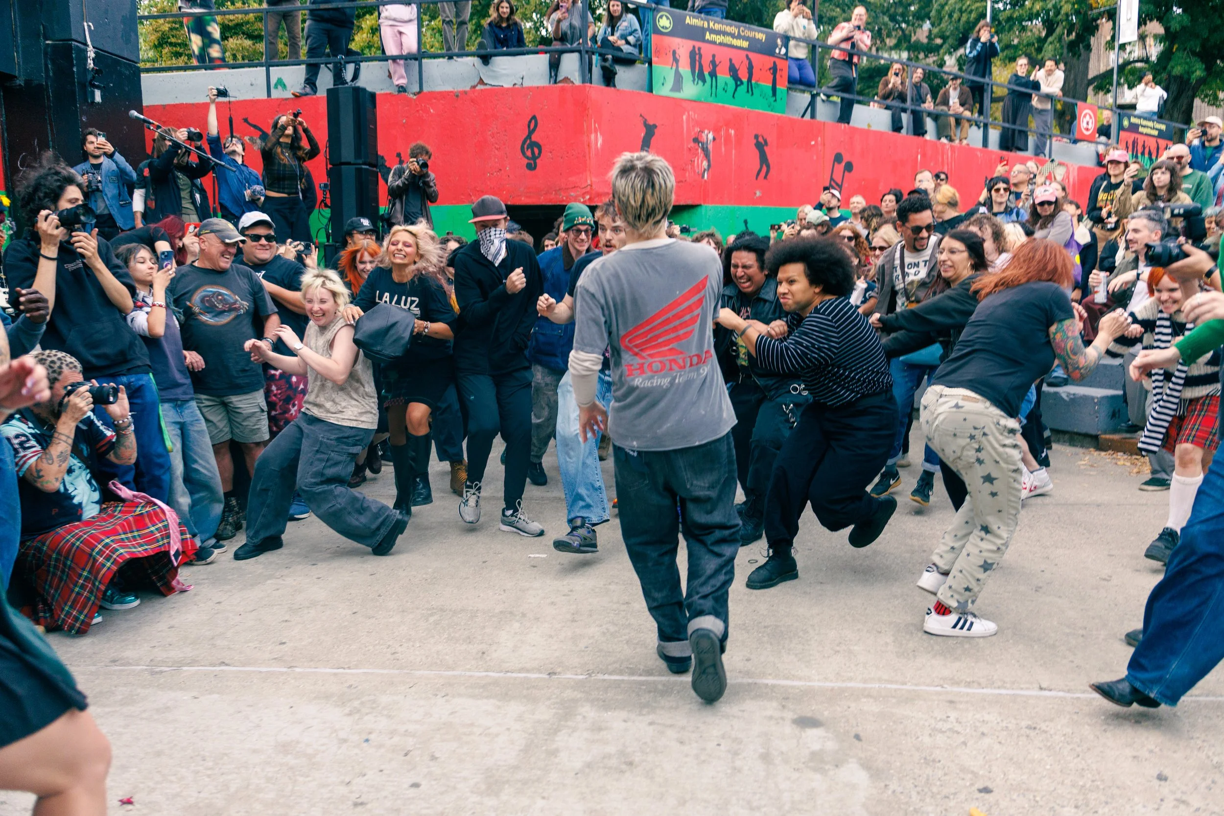 People dancing in a circle at an outdoor event with spectators watching from an elevated platform. The scene is lively and colorful, with some attendees taking photos.