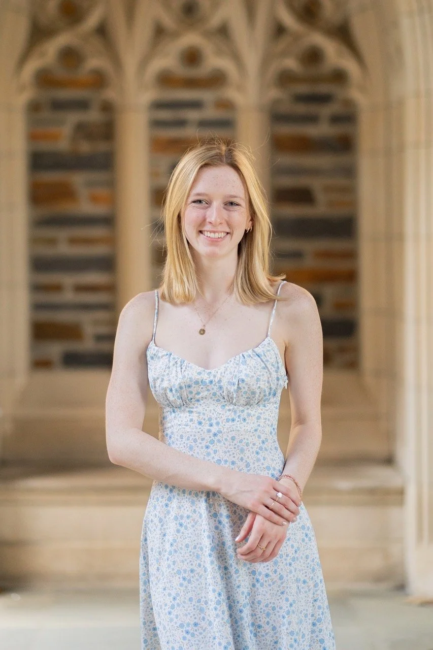 A young woman with blonde hair smiling, wearing a light blue floral dress, standing indoors with a background of arched windows and a stone wall.