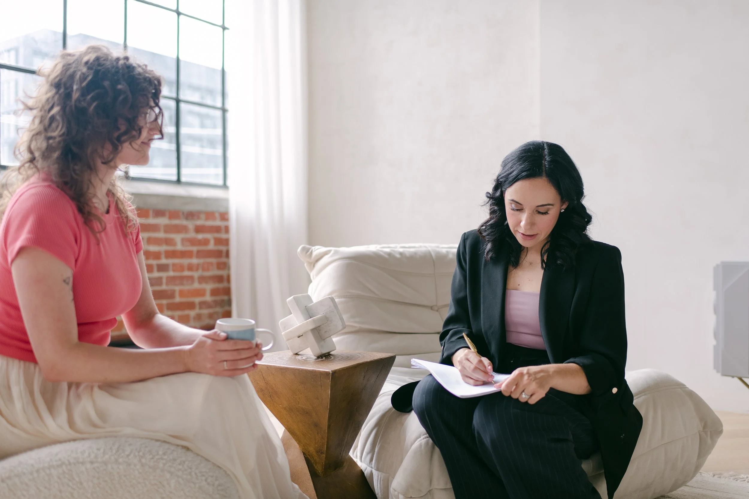 Two women seated indoors, one wearing a pink shirt, holding a mug, and the other in a black blazer, writing on paper. Bright, modern room with large windows and a brick wall.
