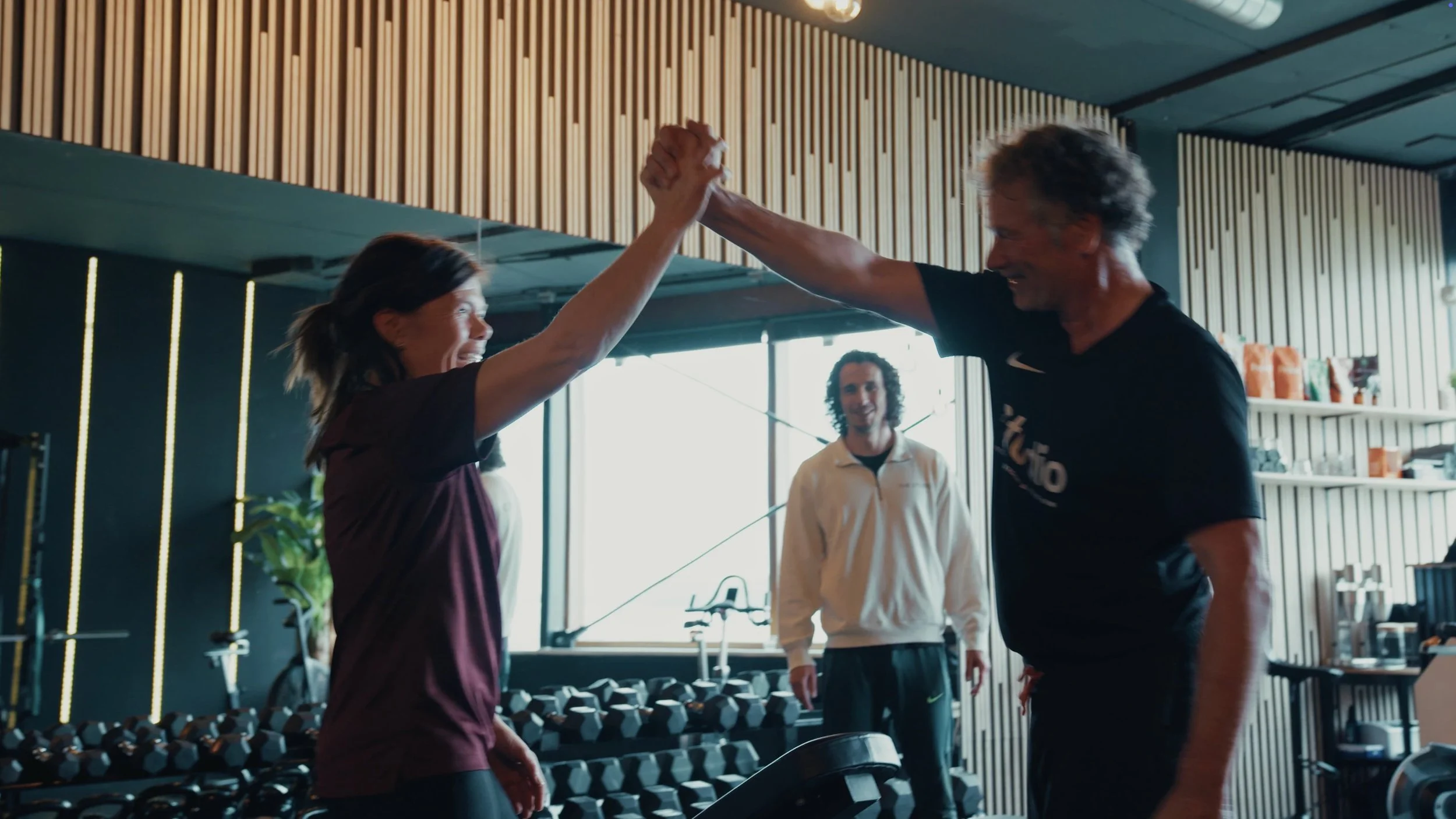Twee mensen in een sportschool die elkaar high-five geven, terwijl een andere persoon in de achtergrond toekijkt.