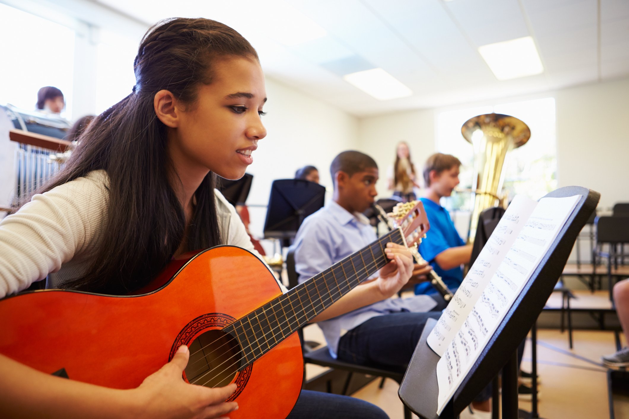 Students playing musical instruments in a classroom, including guitar and clarinet, with sheet music on stands.