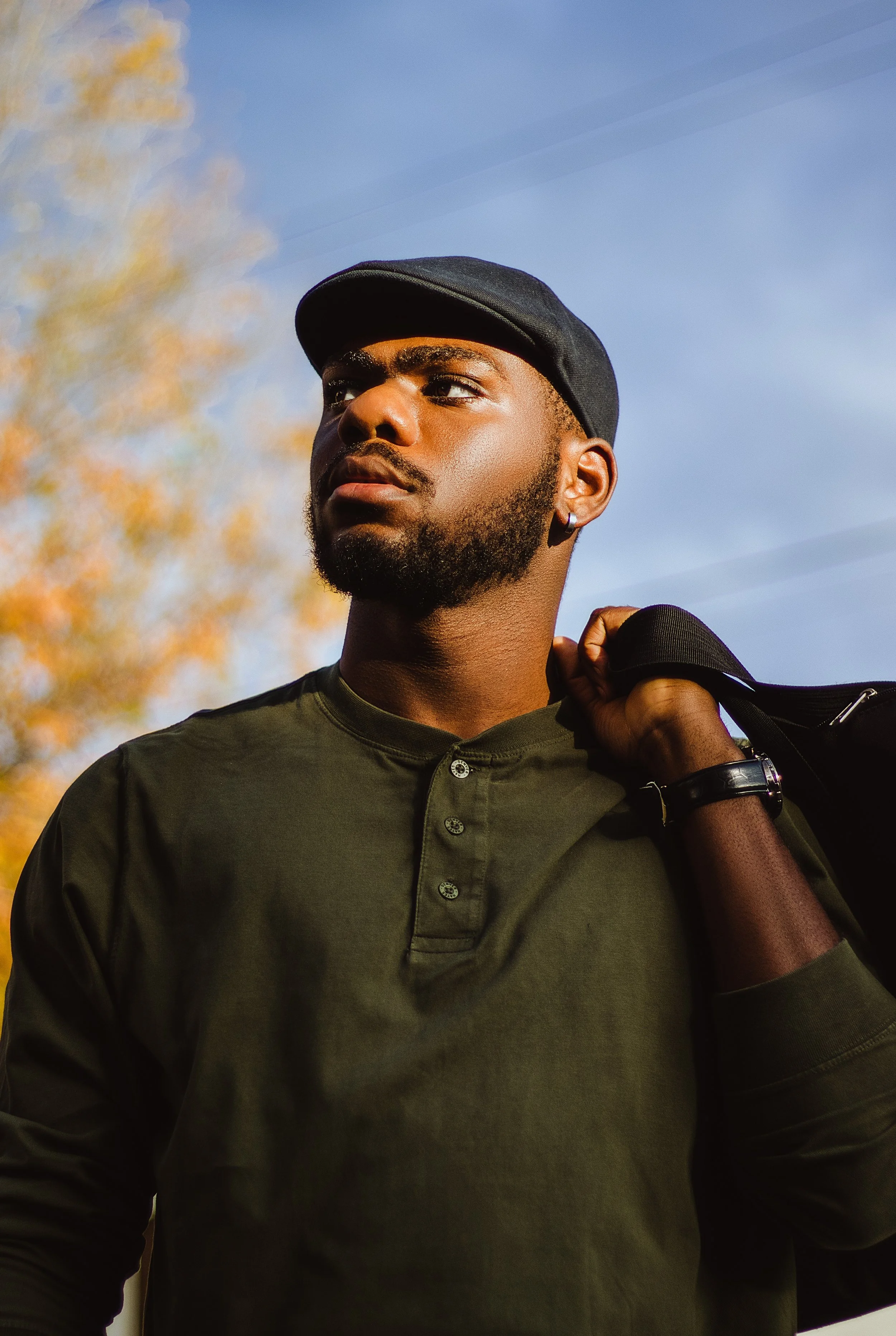 A young man outdoors wearing a black cap, olive green shirt, and holding a bag over his shoulder, with autumn leaves and a clear blue sky in the background.