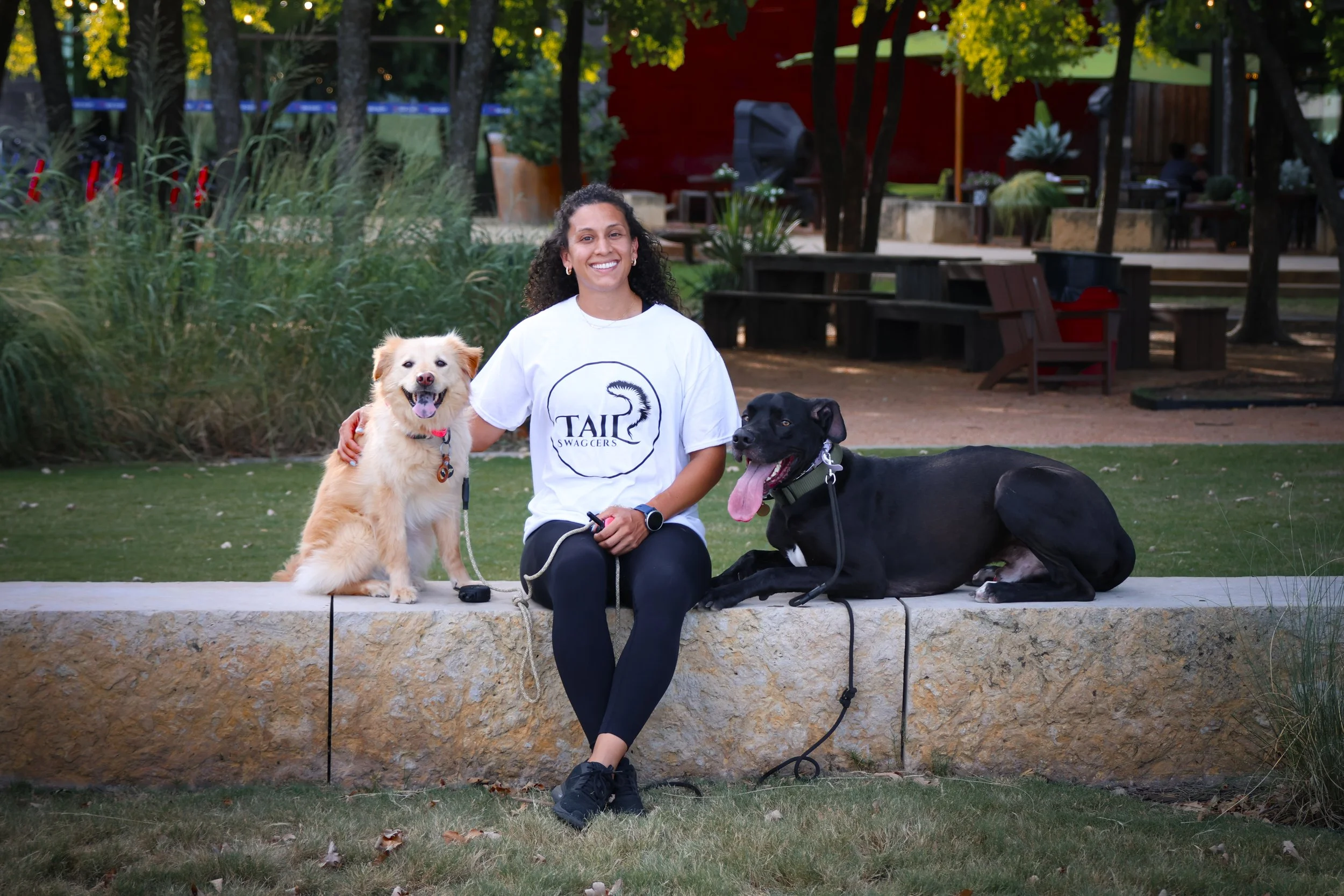 A woman smiling and sitting on a stone ledge outdoors with two dogs, a fluffy light-colored dog and a black dog with a tongue out, in a park or garden setting.