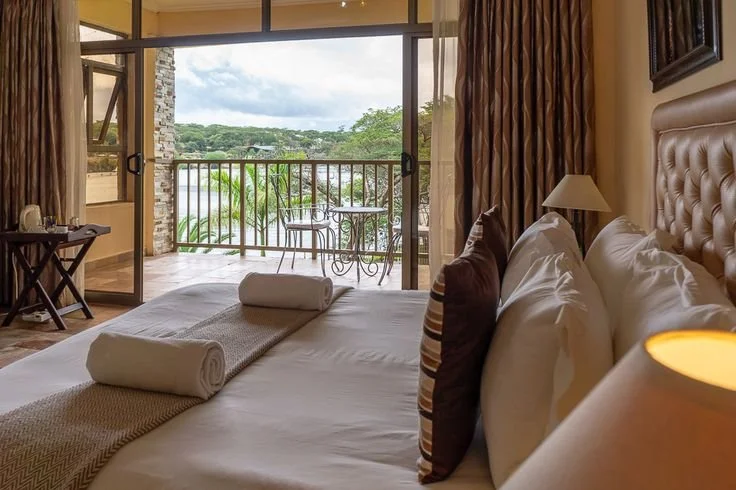 Hotel room with a bed, pillows, and a balcony with outdoor furniture overlooking a lush landscape.