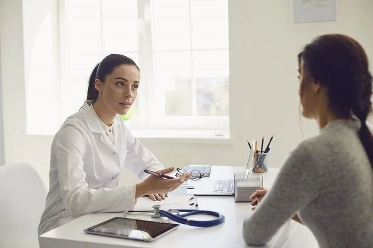 A woman in a white lab coat holding glasses and a clipboard speaking with a patient at a doctor's office.