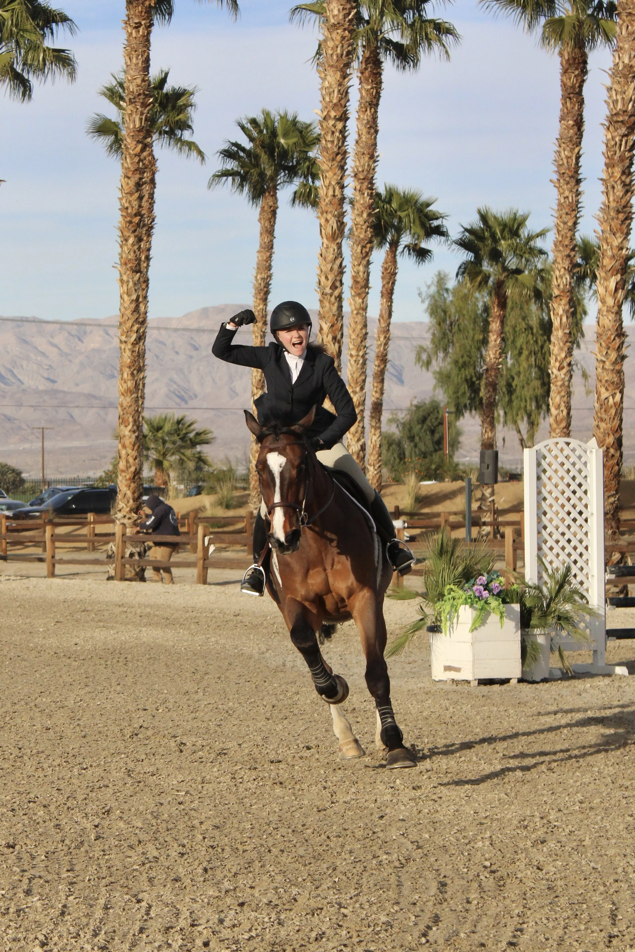 A woman riding a horse in an outdoor equestrian event, raising her right arm in celebration, with palm trees and mountains in the background.