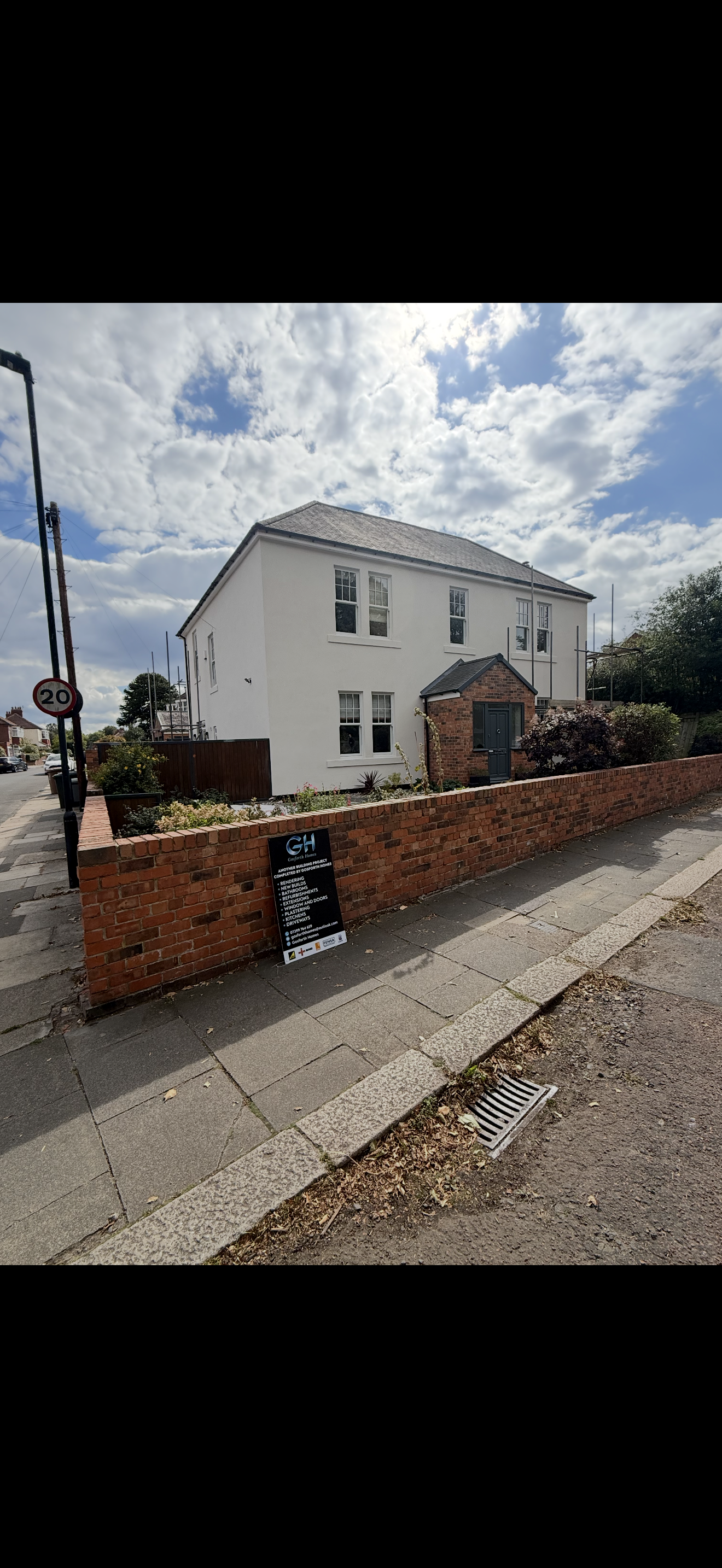 A white two-story house with a brick extension and black front door, surrounded by a low brick wall and a garden, under a partly cloudy sky.