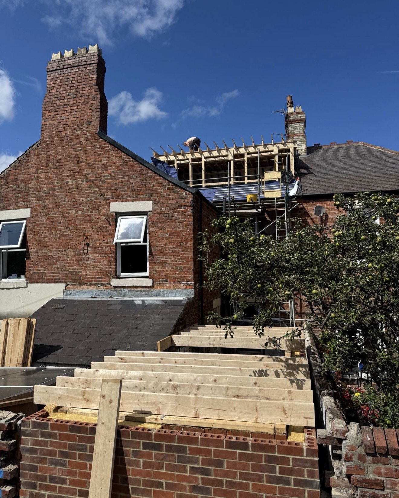 A house with a red brick exterior undergoing construction. The roof is partially removed, with a worker on the roof framing a new section. Construction materials, including wooden planks and scaffolding, are visible in the foreground. A tree with green leaves and fruit is in the yard, with a clear blue sky overhead.