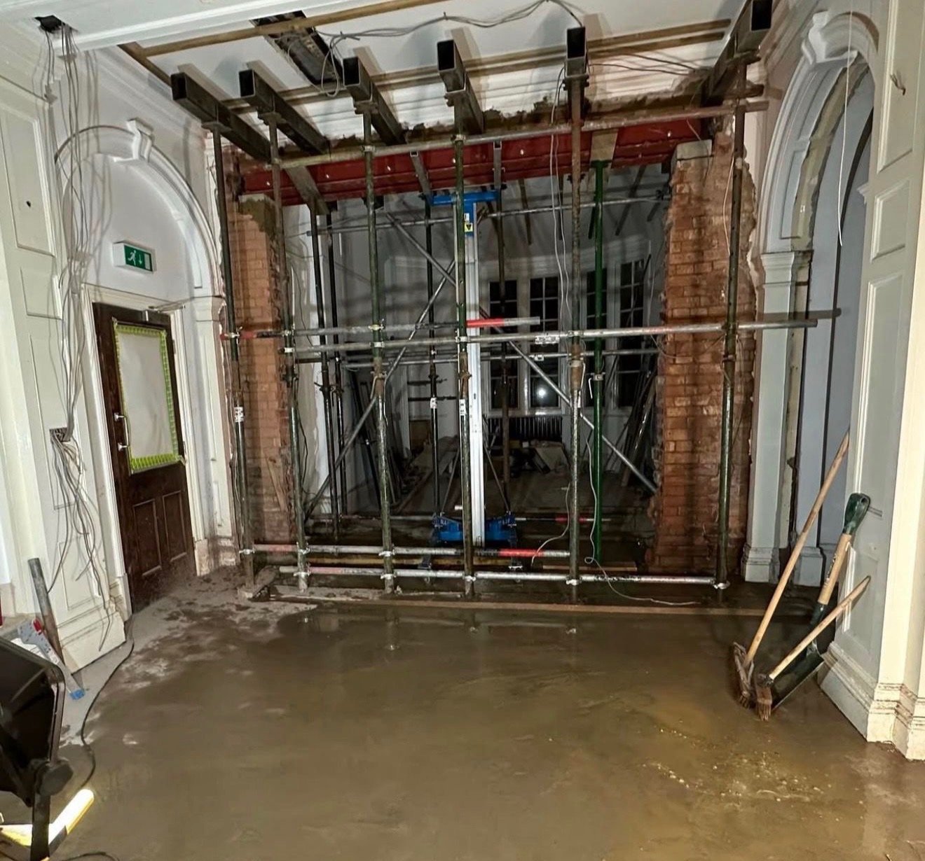 Construction scaffolding set up inside a room with a partially removed wall and exposed brick. The floor appears wet or freshly poured, and there are construction tools leaning against the wall.