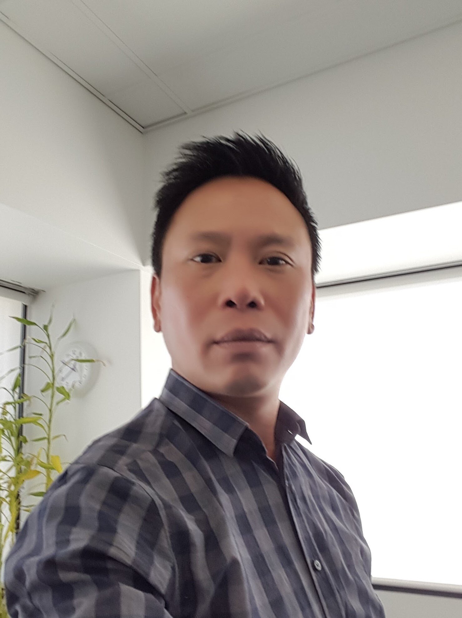 A man taking a selfie inside a modern office. He has short black hair, is wearing a dark plaid shirt, and is standing near a window with bright light behind him. There is a potted plant and a clock on the wall in the background.