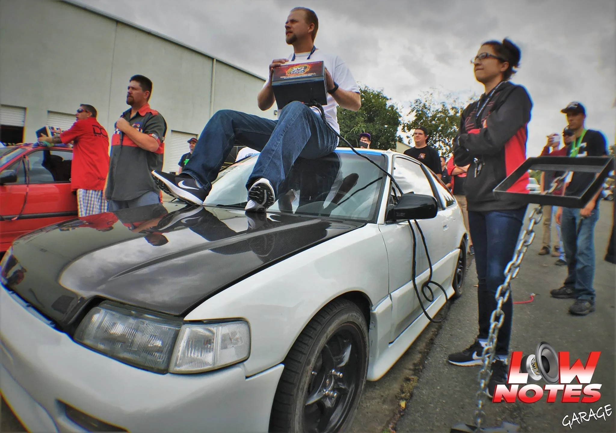 A man sitting on the hood of a white car at a racing event, holding a box, surrounded by people, with a cloudy sky overhead and a chain barrier in the foreground.